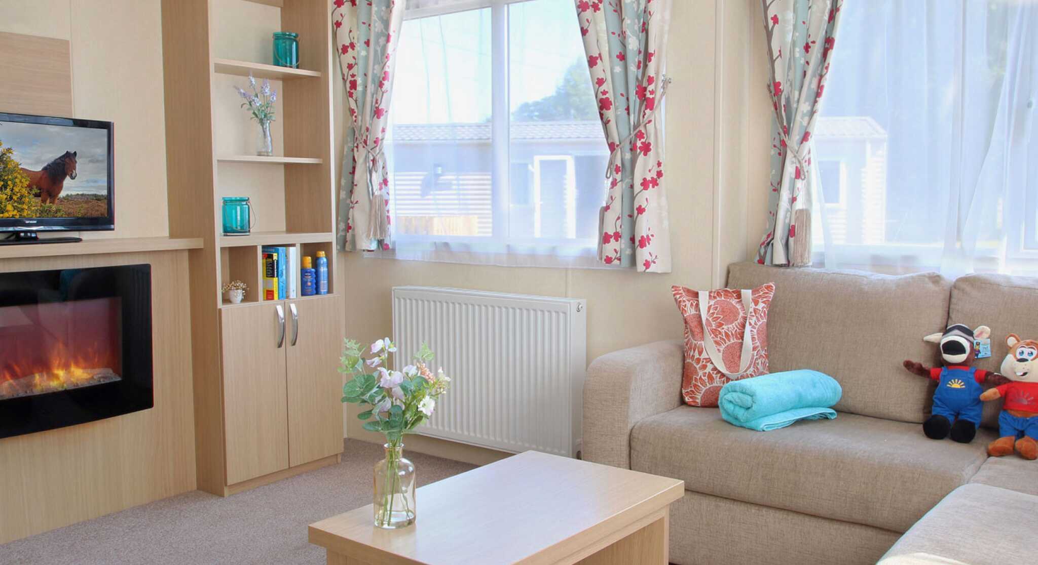 A cozy living room featuring a beige sofa, a wooden coffee table, and a decorative vase with flowers. There are colorful curtains with floral patterns, a television on a shelf, and a faux fireplace below it. A blue towel and stuffed toys are also on the sofa.