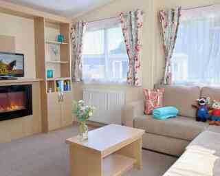 A cozy living room featuring a beige sofa, a wooden coffee table, and a decorative vase with flowers. There are colorful curtains with floral patterns, a television on a shelf, and a faux fireplace below it. A blue towel and stuffed toys are also on the sofa.