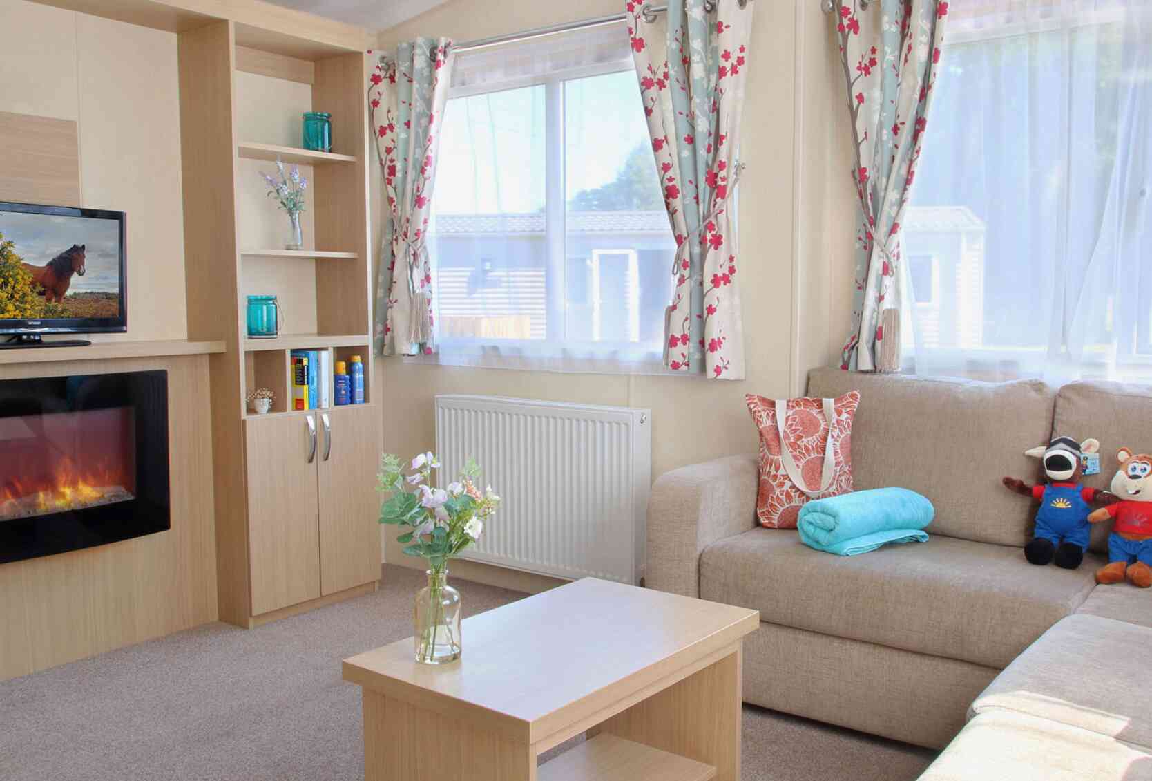 A cozy living room featuring a beige sofa, a wooden coffee table, and a decorative vase with flowers. There are colorful curtains with floral patterns, a television on a shelf, and a faux fireplace below it. A blue towel and stuffed toys are also on the sofa.