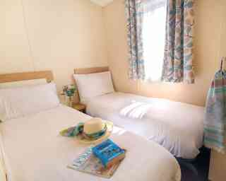 A cozy bedroom with two single beds, one with a sun hat and a beach towel on it. A stack of magazines is placed on a bedside table. A window with patterned curtains allows natural light to brighten the room.