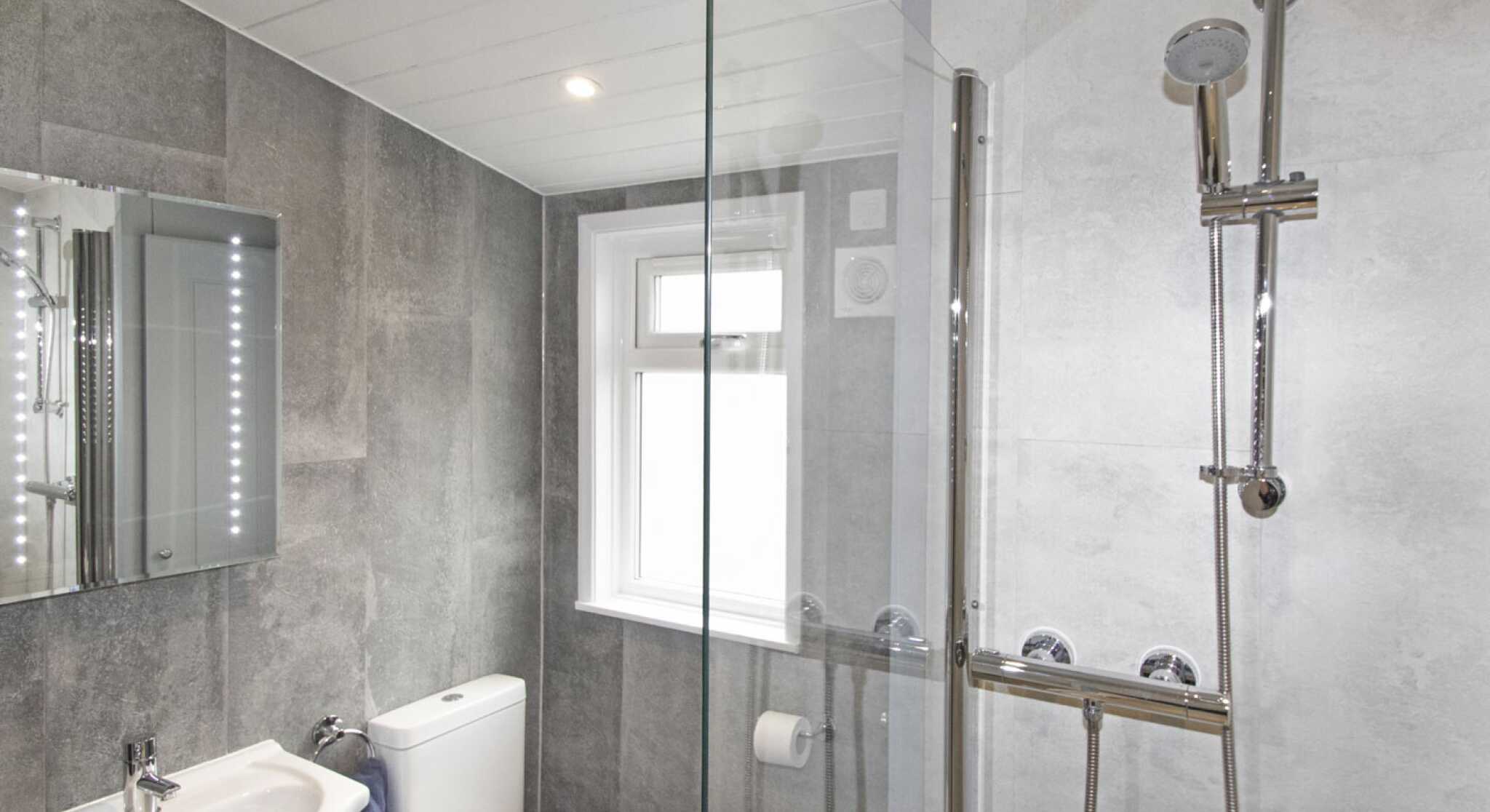 Modern bathroom featuring a glass shower enclosure, sleek silver fixtures, a white toilet, and a compact sink with a mirror. Gray tiled walls and flooring create a contemporary look.