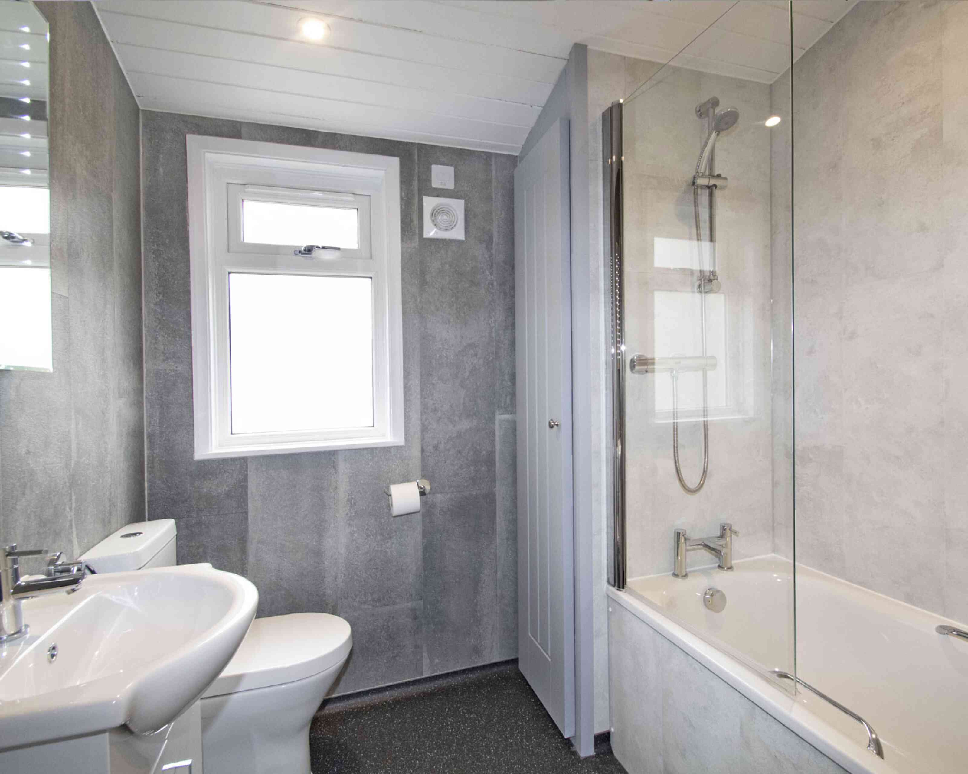 A modern bathroom featuring a bathtub with a glass shower screen, a toilet, and a sink. The walls have a gray finish, and there is a window allowing natural light.