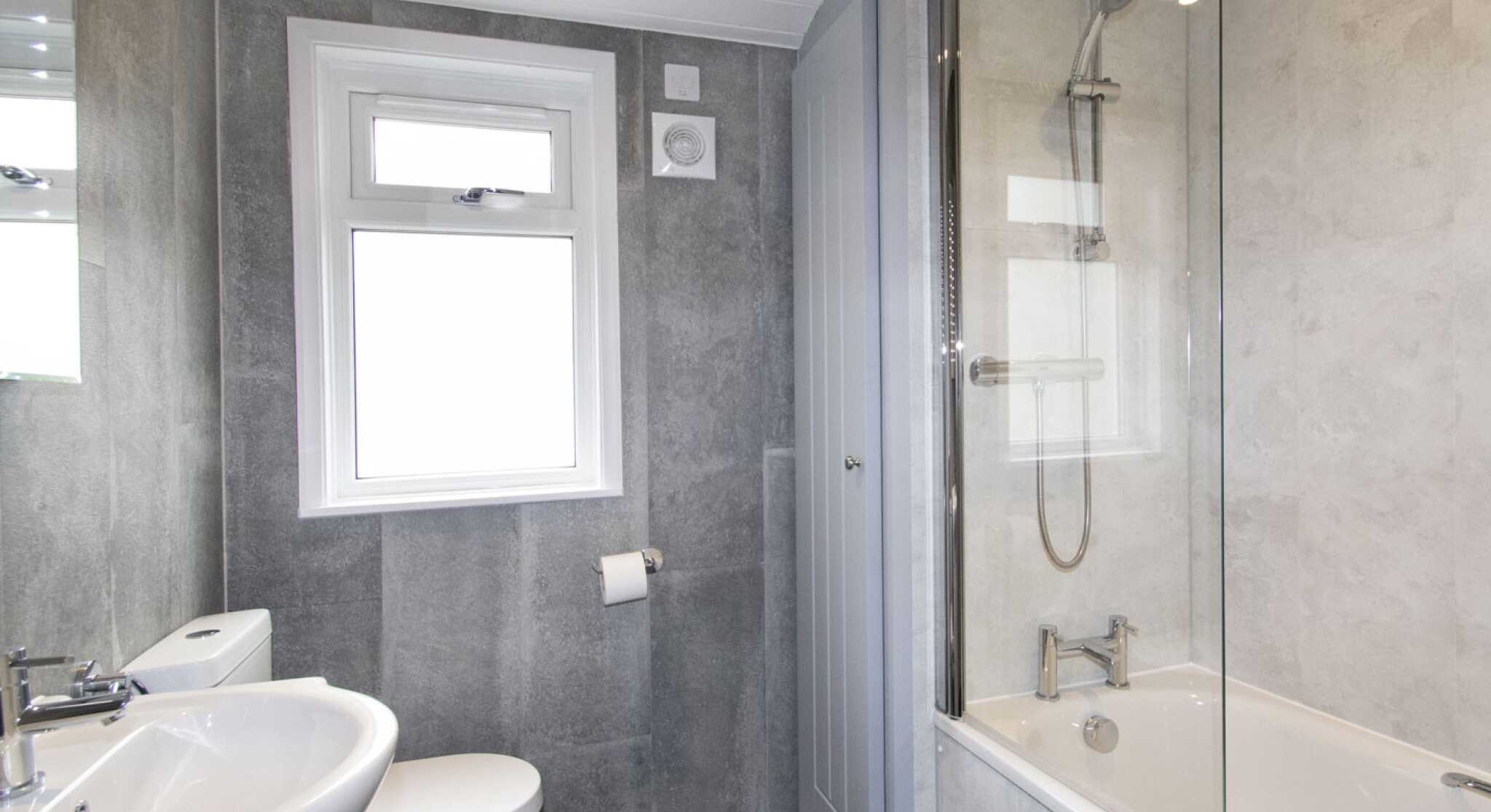 A modern bathroom featuring a bathtub with a glass shower screen, a toilet, and a sink. The walls have a gray finish, and there is a window allowing natural light.