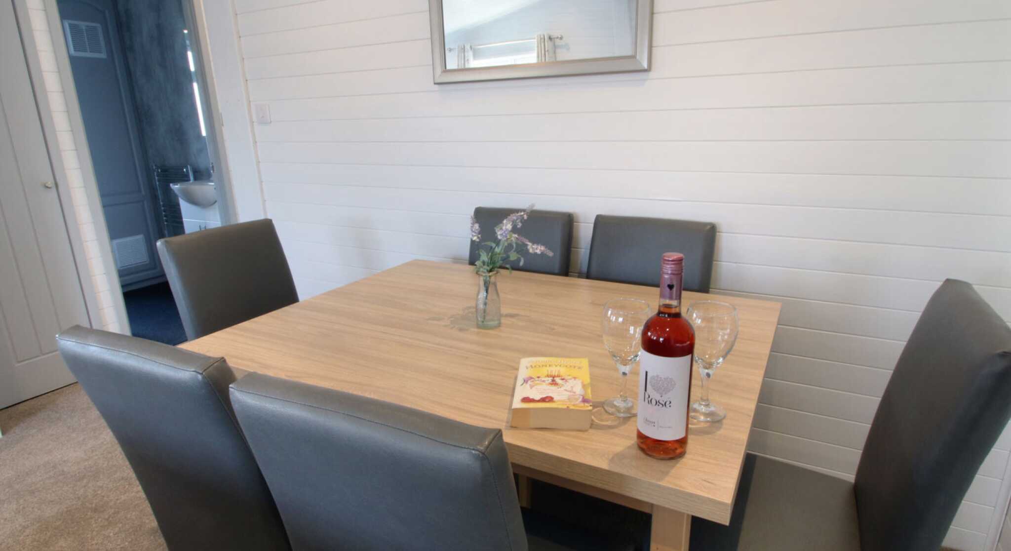 A modern dining table with four gray chairs, featuring a small vase with flowers and a bottle of rosé wine alongside two empty glasses and a book. A mirror hangs on the wall, and a doorway to another room is visible in the background.