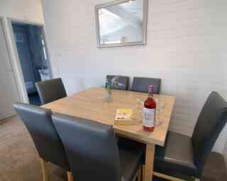 A modern dining table with four gray chairs, featuring a small vase with flowers and a bottle of rosé wine alongside two empty glasses and a book. A mirror hangs on the wall, and a doorway to another room is visible in the background.