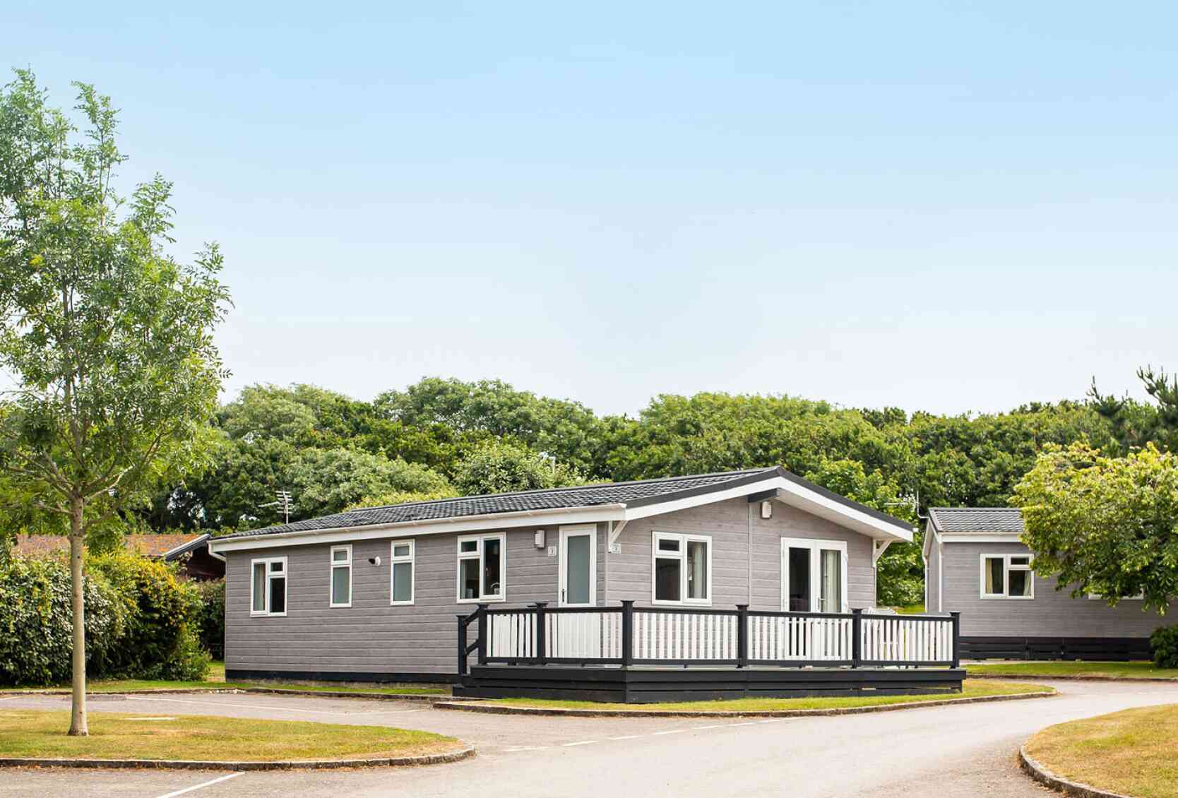 A modern single-story gray mobile home with a black railing deck is situated in a green area with trees. A paved road curves around the home, leading to other similar structures in the background. Clear blue sky is overhead.
