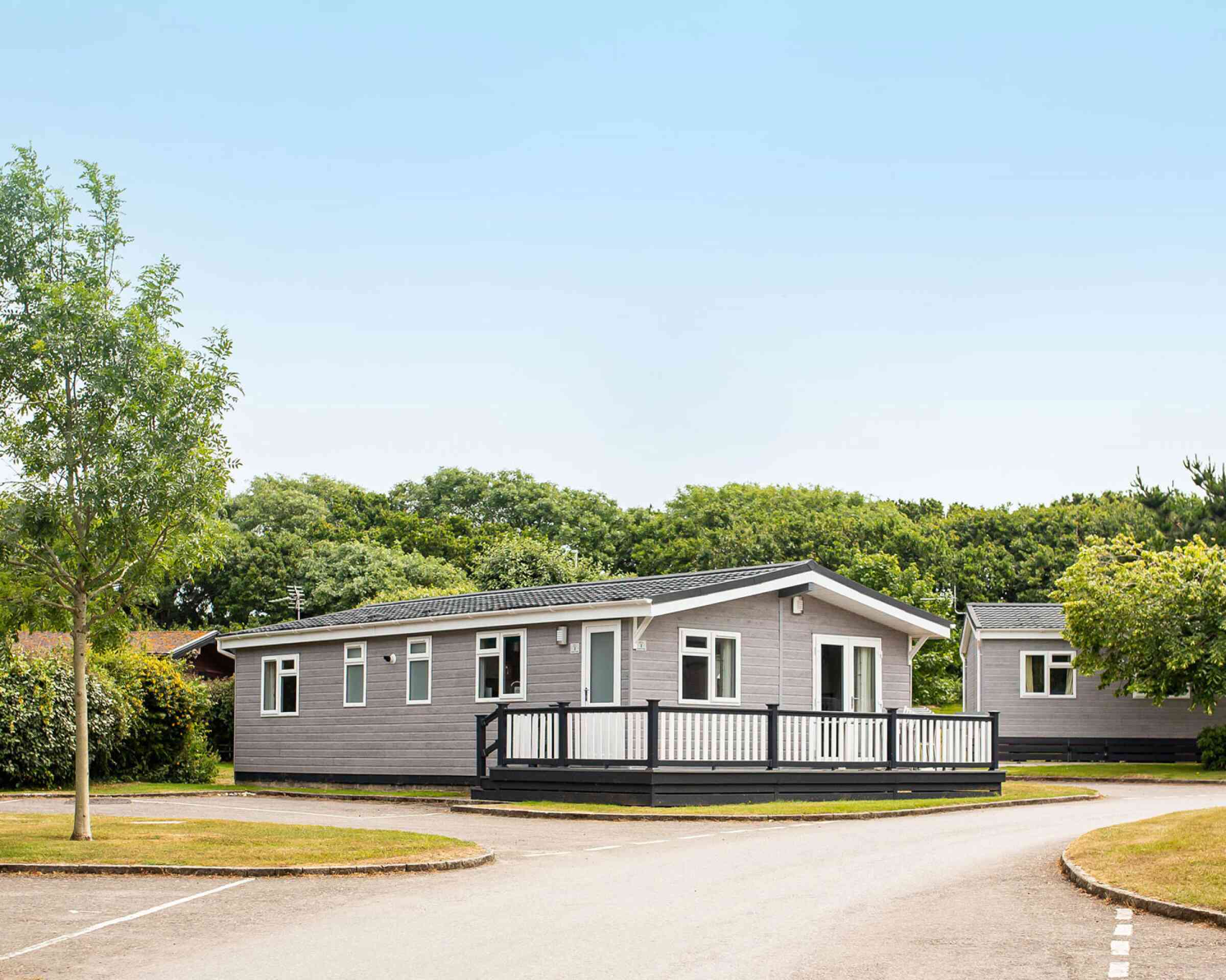 A modern single-story gray mobile home with a black railing deck is situated in a green area with trees. A paved road curves around the home, leading to other similar structures in the background. Clear blue sky is overhead.