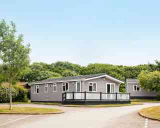 A modern single-story gray mobile home with a black railing deck is situated in a green area with trees. A paved road curves around the home, leading to other similar structures in the background. Clear blue sky is overhead.