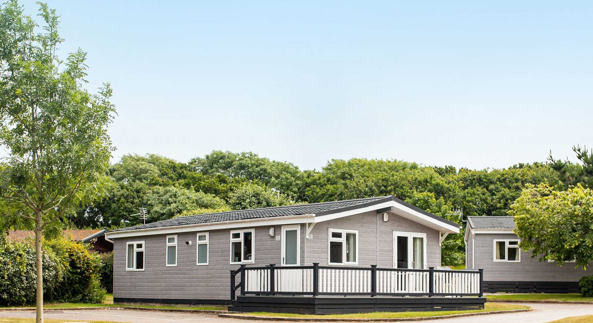 A modern single-story gray mobile home with a black railing deck is situated in a green area with trees. A paved road curves around the home, leading to other similar structures in the background. Clear blue sky is overhead.