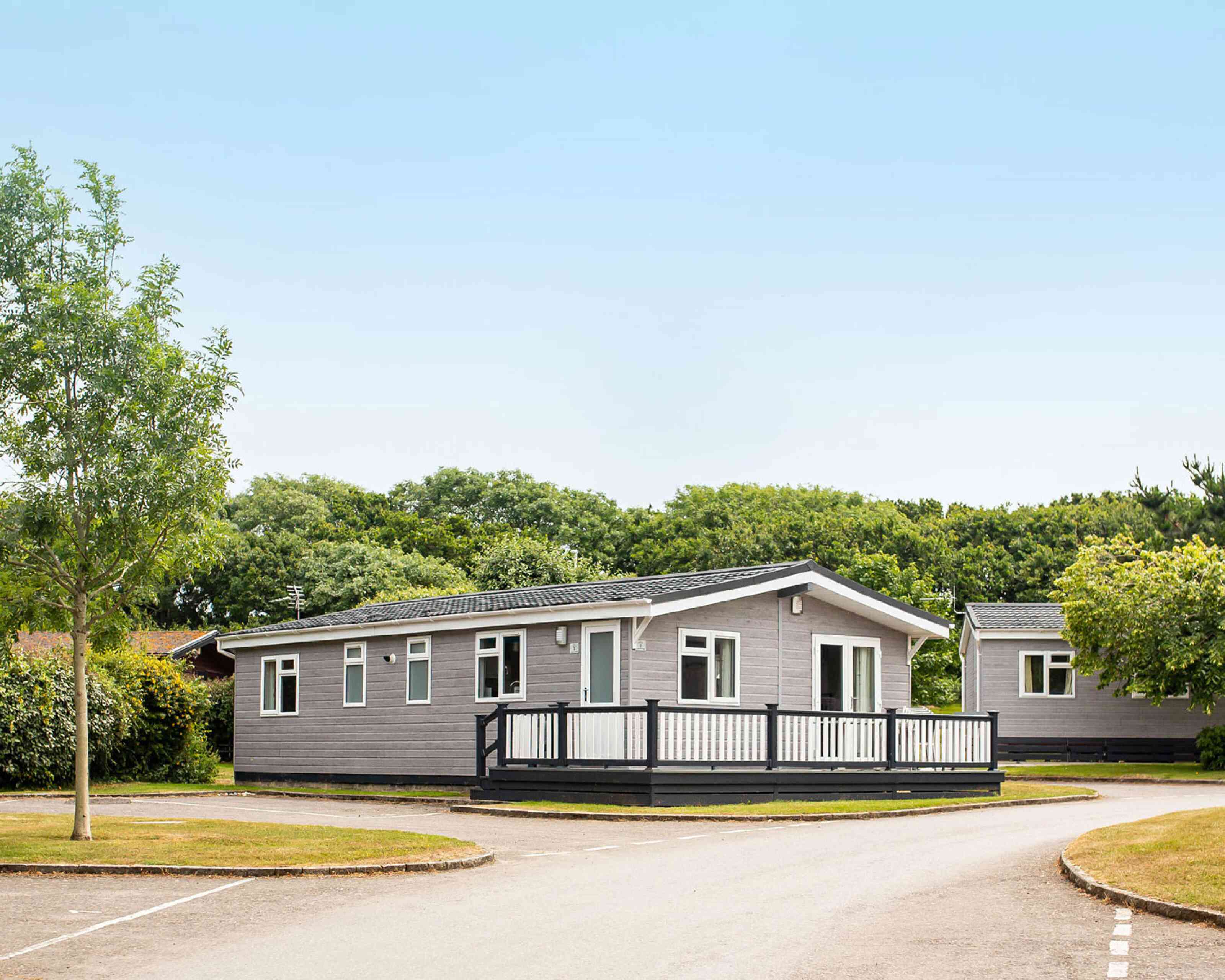 A modern single-story gray mobile home with a black railing deck is situated in a green area with trees. A paved road curves around the home, leading to other similar structures in the background. Clear blue sky is overhead.