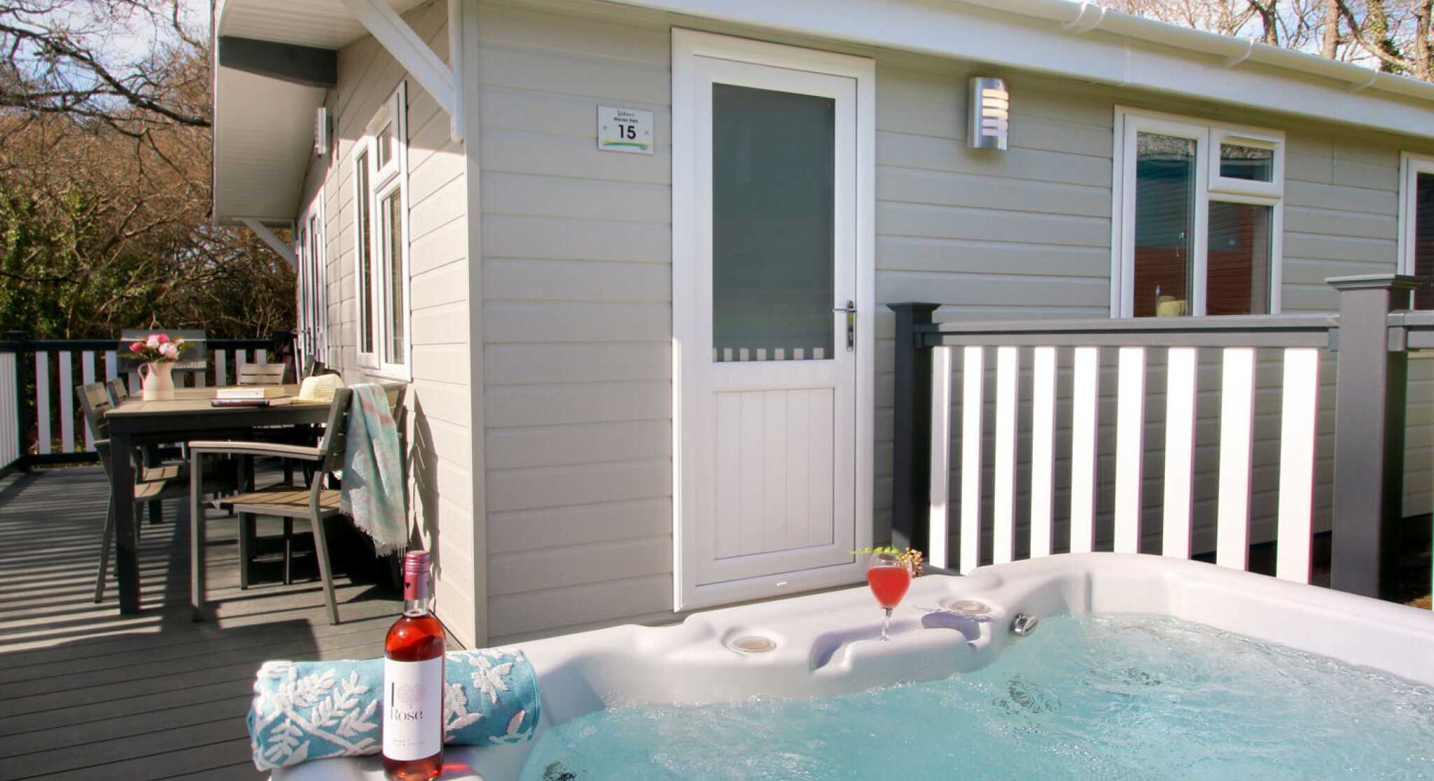 A hot tub filled with water sits on a wooden deck next to a bottle of rosé wine and a glass with a cocktail. In the background, a cozy cabin with a white door and windows is visible, surrounded by trees.