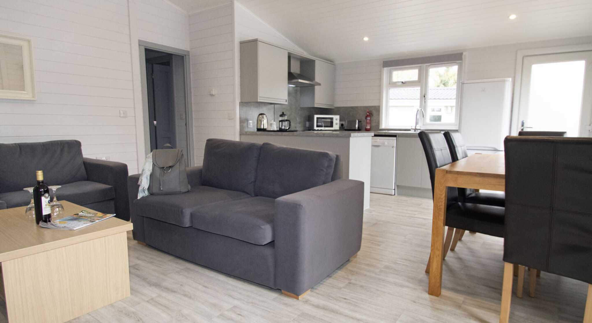 A modern, open-plan living space with a gray sofa and a wooden coffee table in the foreground. The kitchen area features sleek cabinets and appliances, while a dining table with dark chairs is visible in the background. Bright, natural light enters through large windows.
