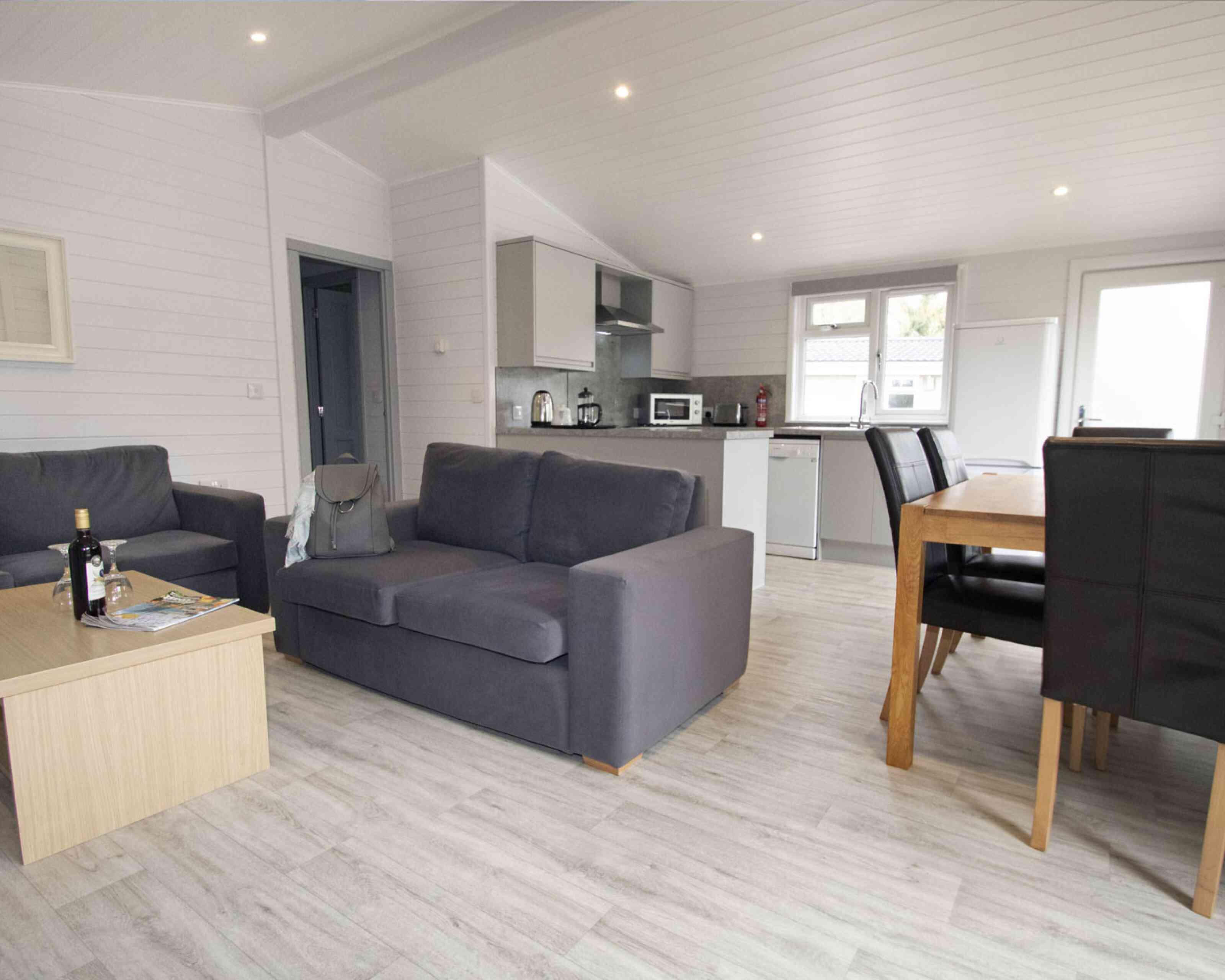 A modern, open-plan living space with a gray sofa and a wooden coffee table in the foreground. The kitchen area features sleek cabinets and appliances, while a dining table with dark chairs is visible in the background. Bright, natural light enters through large windows.