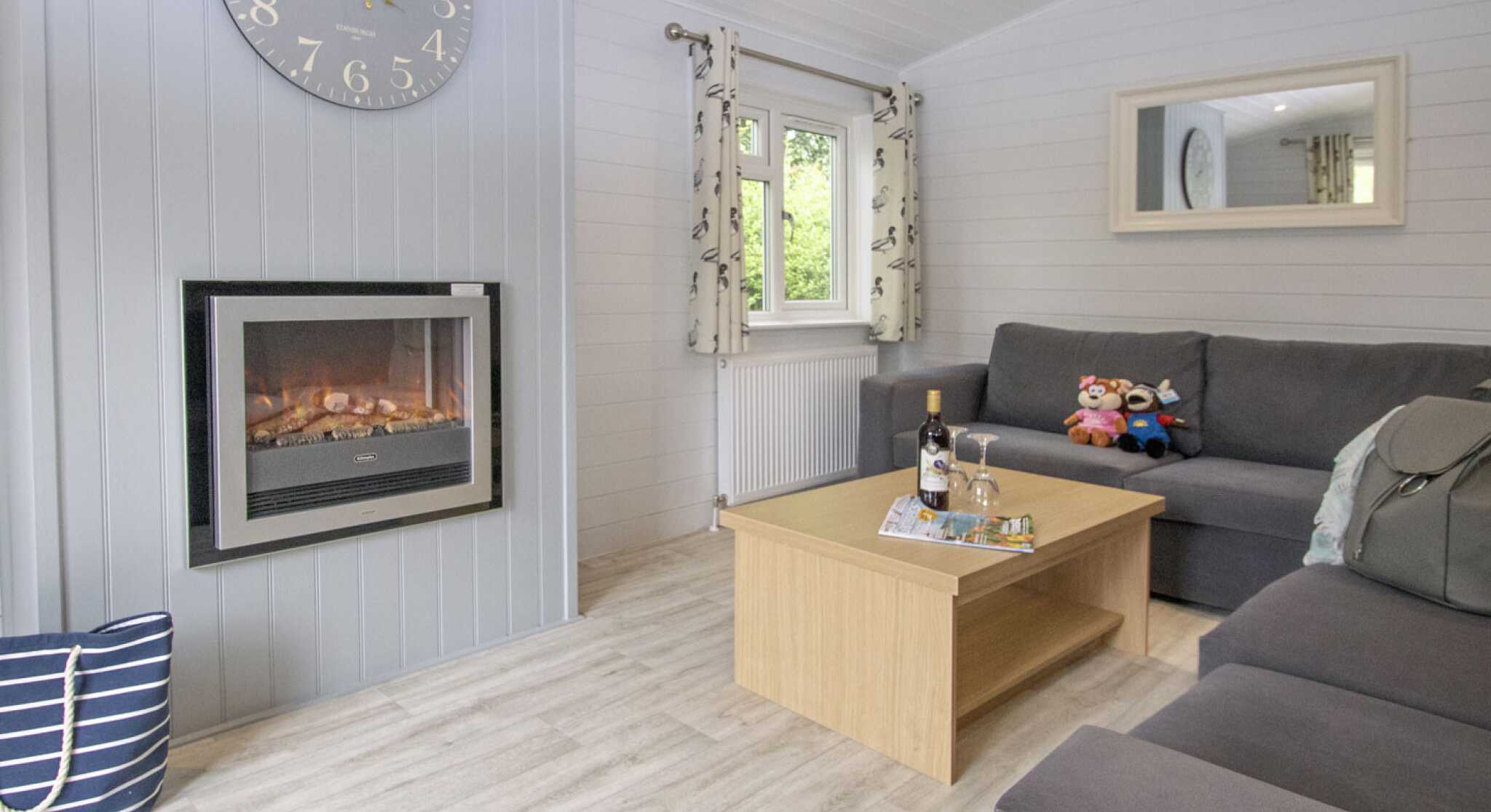 Cozy living room featuring a gray sofa, a wooden coffee table with decorative items, and a stylish clock on the wall. A modern electric fireplace adds warmth, while large windows let in natural light. A plush toy and a cushion are on the sofa.