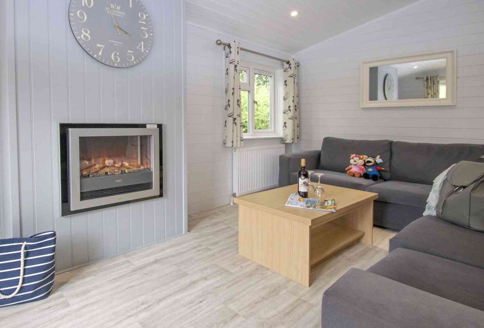 Cozy living room featuring a gray sofa, a wooden coffee table with decorative items, and a stylish clock on the wall. A modern electric fireplace adds warmth, while large windows let in natural light. A plush toy and a cushion are on the sofa.