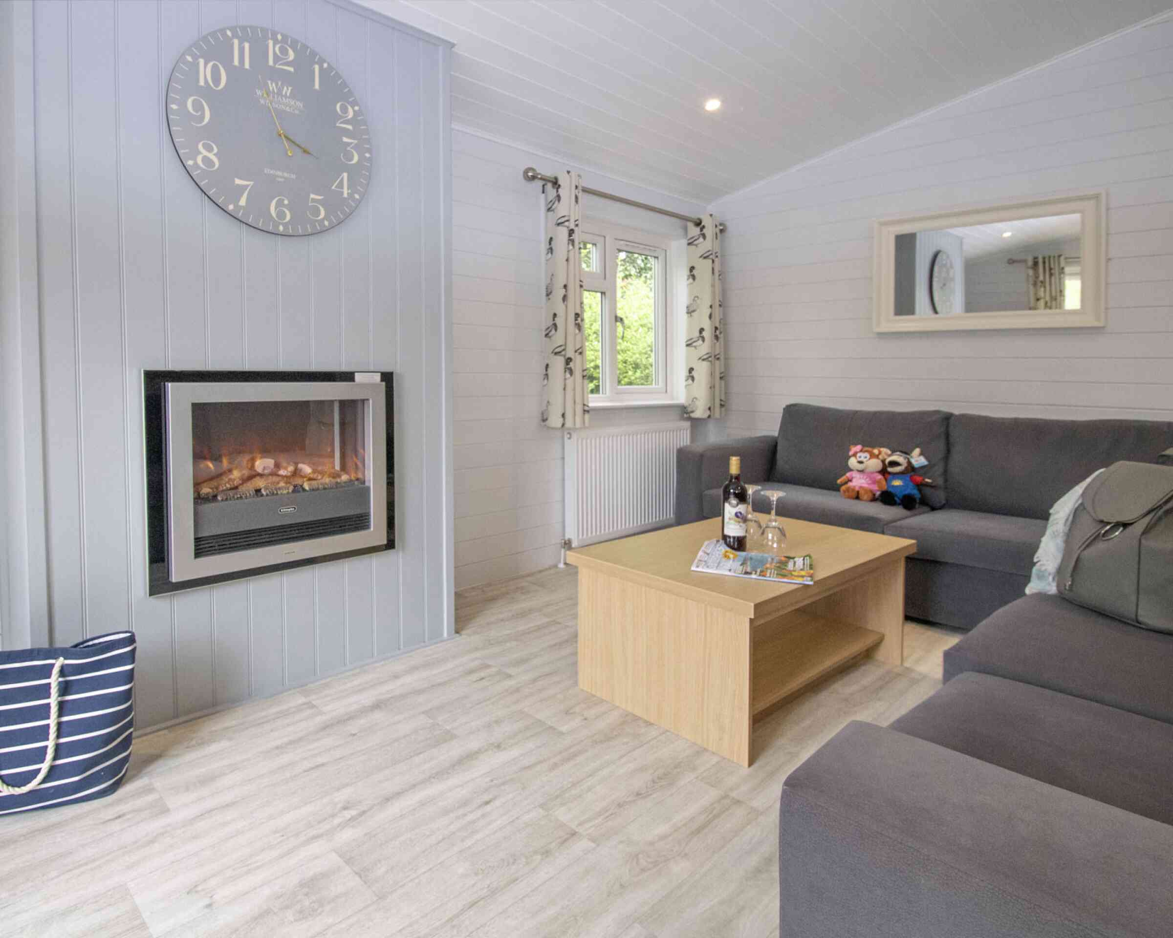 Cozy living room featuring a gray sofa, a wooden coffee table with decorative items, and a stylish clock on the wall. A modern electric fireplace adds warmth, while large windows let in natural light. A plush toy and a cushion are on the sofa.