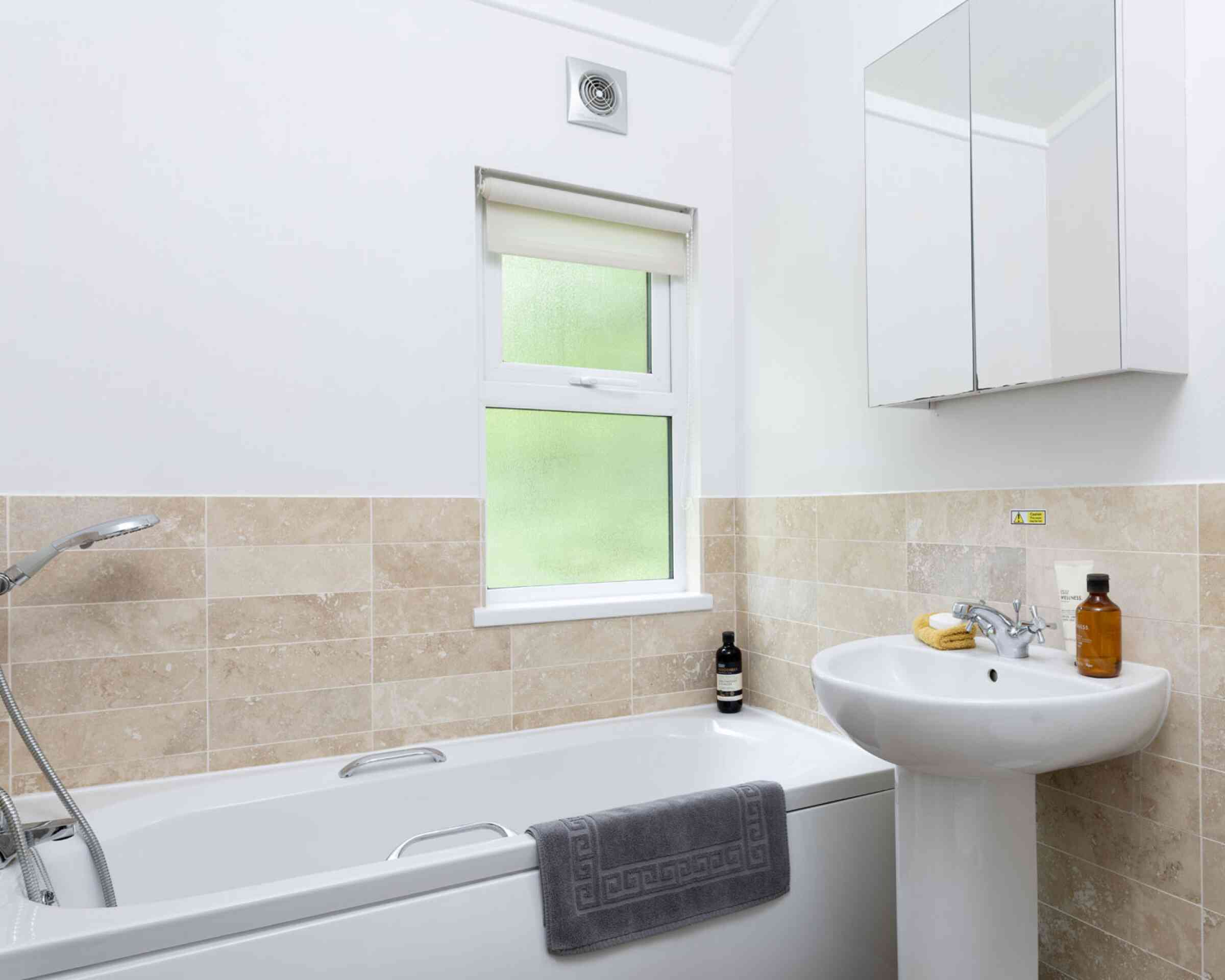 A modern bathroom featuring a white bathtub with a gray towel draped over it, a pedestal sink, and a window with a light-filtering blind. The walls are tiled with beige stone, and a mirror hangs above the sink. Bottles and a banana are on the sink.