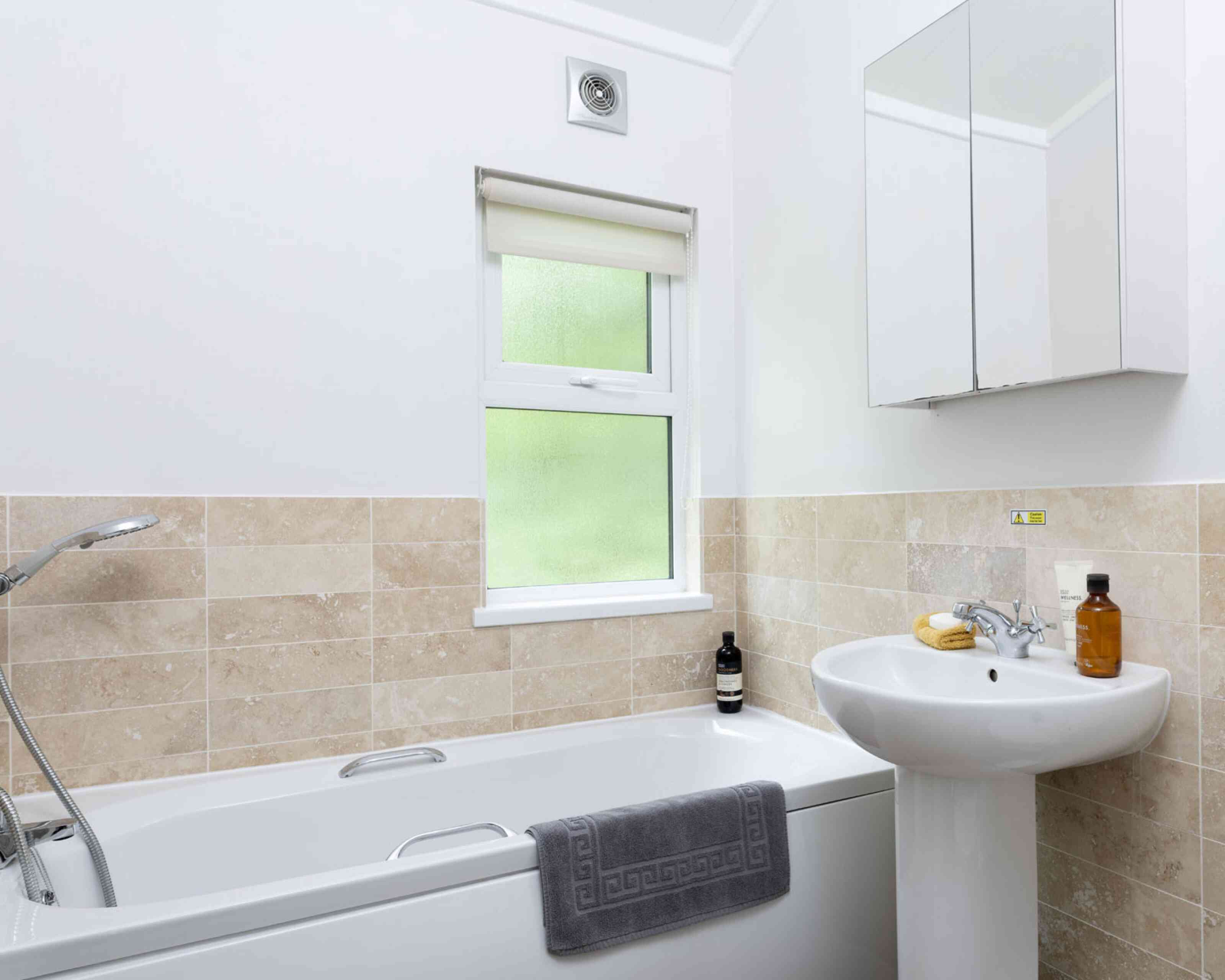 A modern bathroom featuring a white bathtub with a gray towel draped over it, a pedestal sink, and a window with a light-filtering blind. The walls are tiled with beige stone, and a mirror hangs above the sink. Bottles and a banana are on the sink.