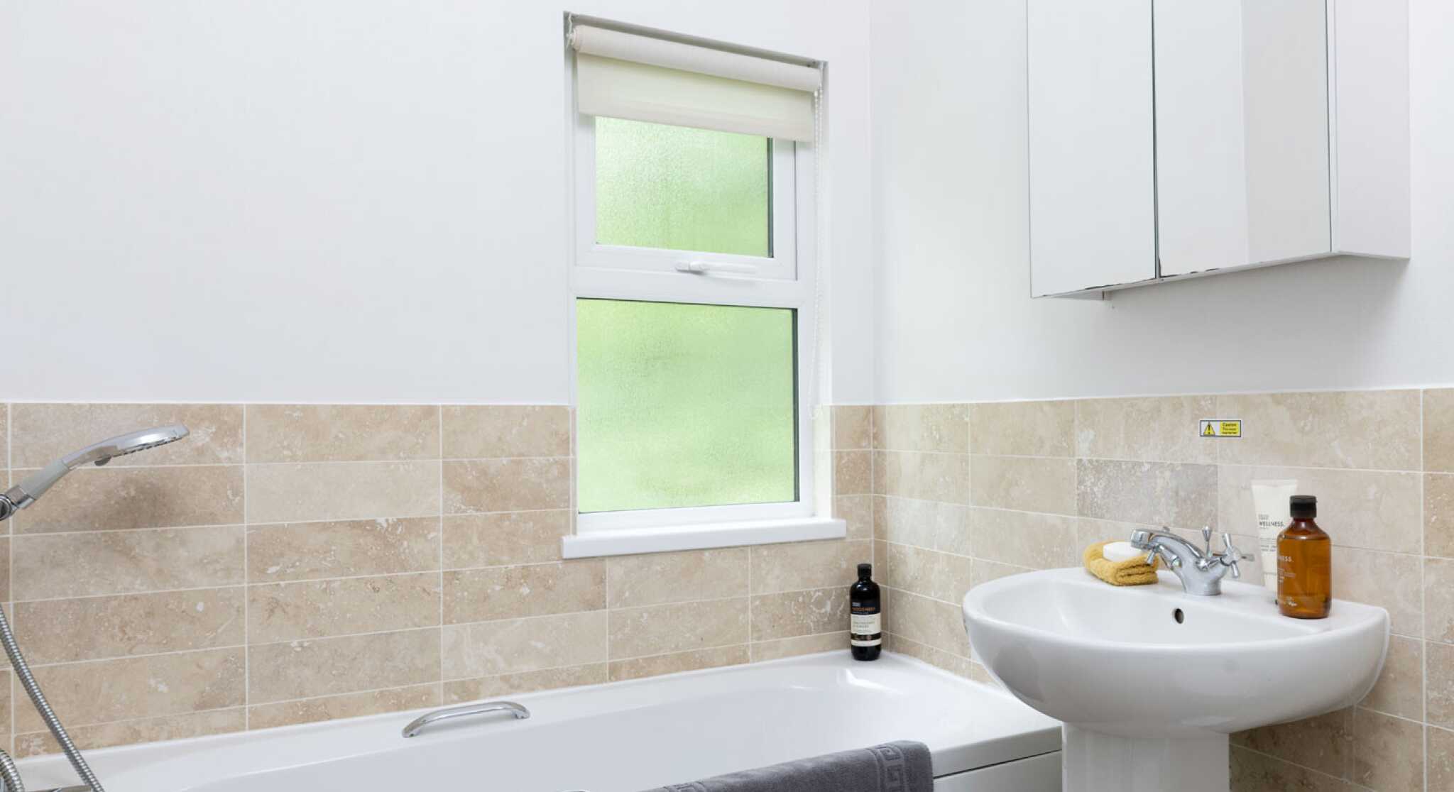 A modern bathroom featuring a white bathtub with a gray towel draped over it, a pedestal sink, and a window with a light-filtering blind. The walls are tiled with beige stone, and a mirror hangs above the sink. Bottles and a banana are on the sink.