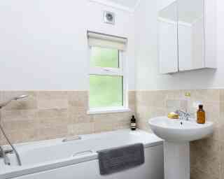 A modern bathroom featuring a white bathtub with a gray towel draped over it, a pedestal sink, and a window with a light-filtering blind. The walls are tiled with beige stone, and a mirror hangs above the sink. Bottles and a banana are on the sink.