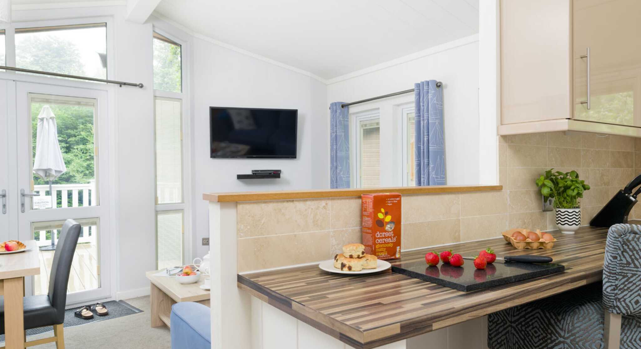 A modern kitchen with a wooden countertop showcases a box of cereal and decorative items. A dining table and chairs are visible in the background, along with a television mounted on the wall and large windows letting in natural light.