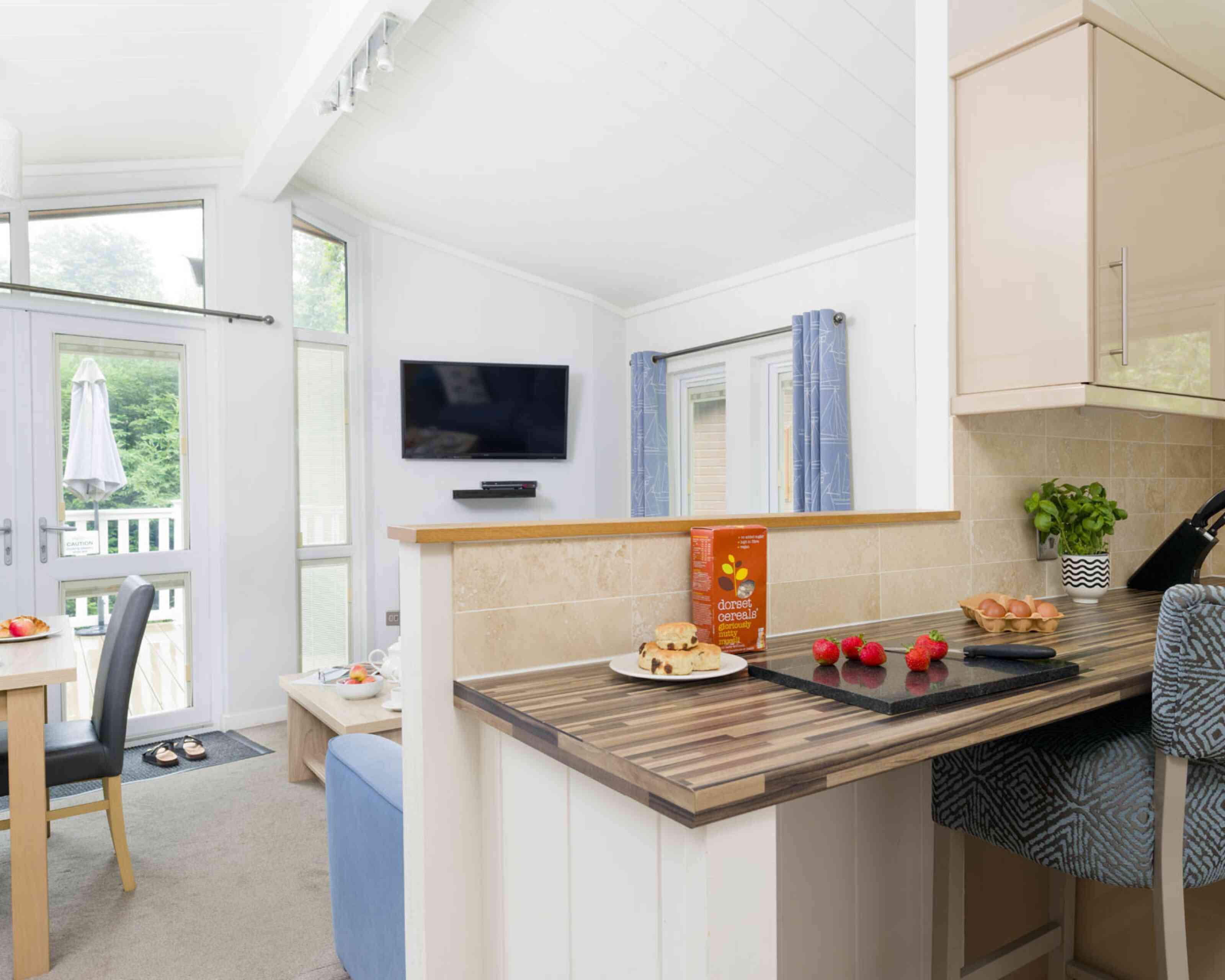 A modern kitchen with a wooden countertop showcases a box of cereal and decorative items. A dining table and chairs are visible in the background, along with a television mounted on the wall and large windows letting in natural light.