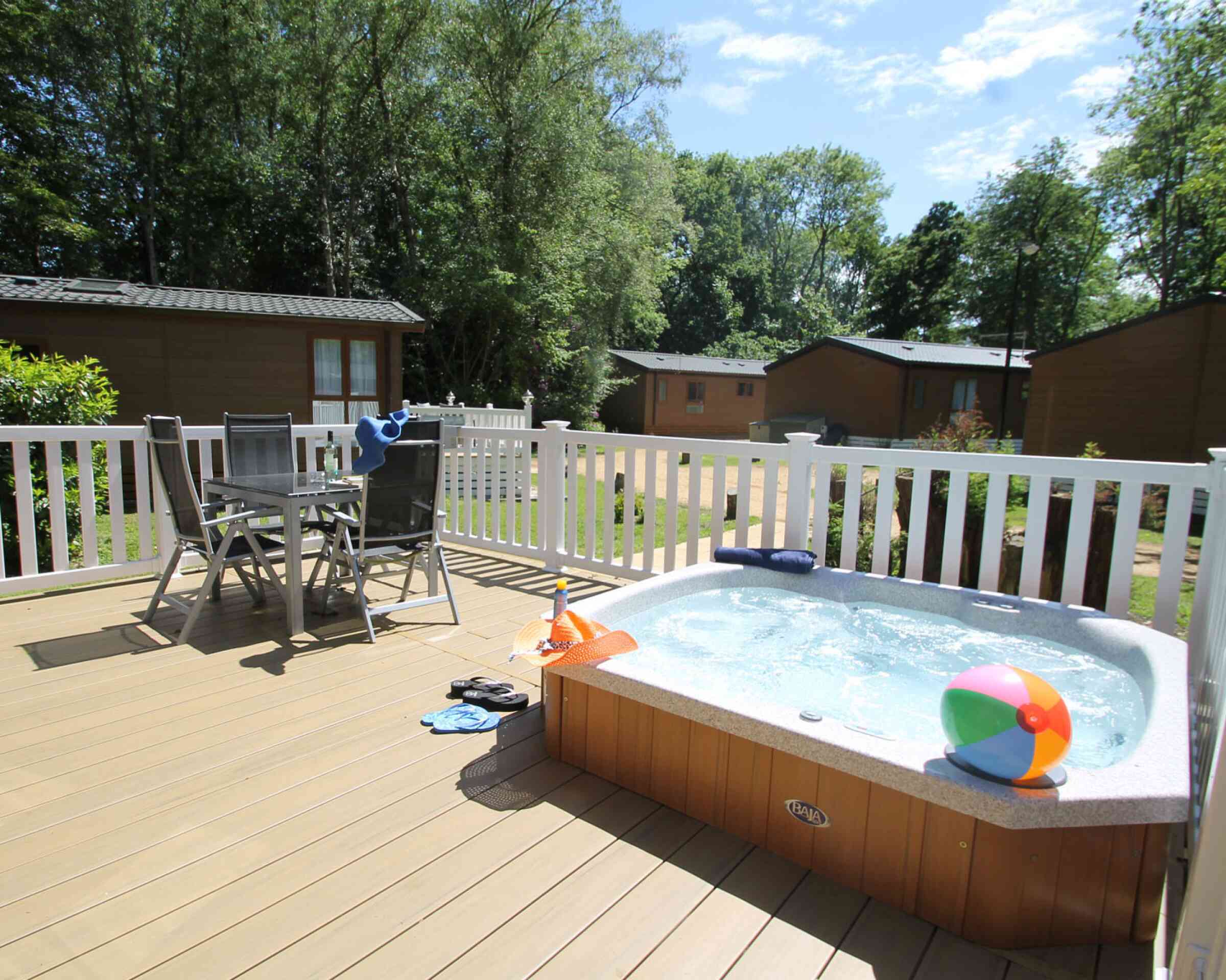 A hot tub on a wooden deck with a beach ball nearby. Two chairs and a small table are set up beside the tub, surrounded by greenery and cabins in the background. The sky is clear with a few clouds.
