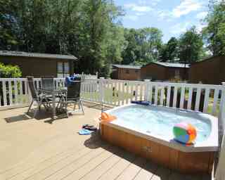 A hot tub on a wooden deck with a beach ball nearby. Two chairs and a small table are set up beside the tub, surrounded by greenery and cabins in the background. The sky is clear with a few clouds.