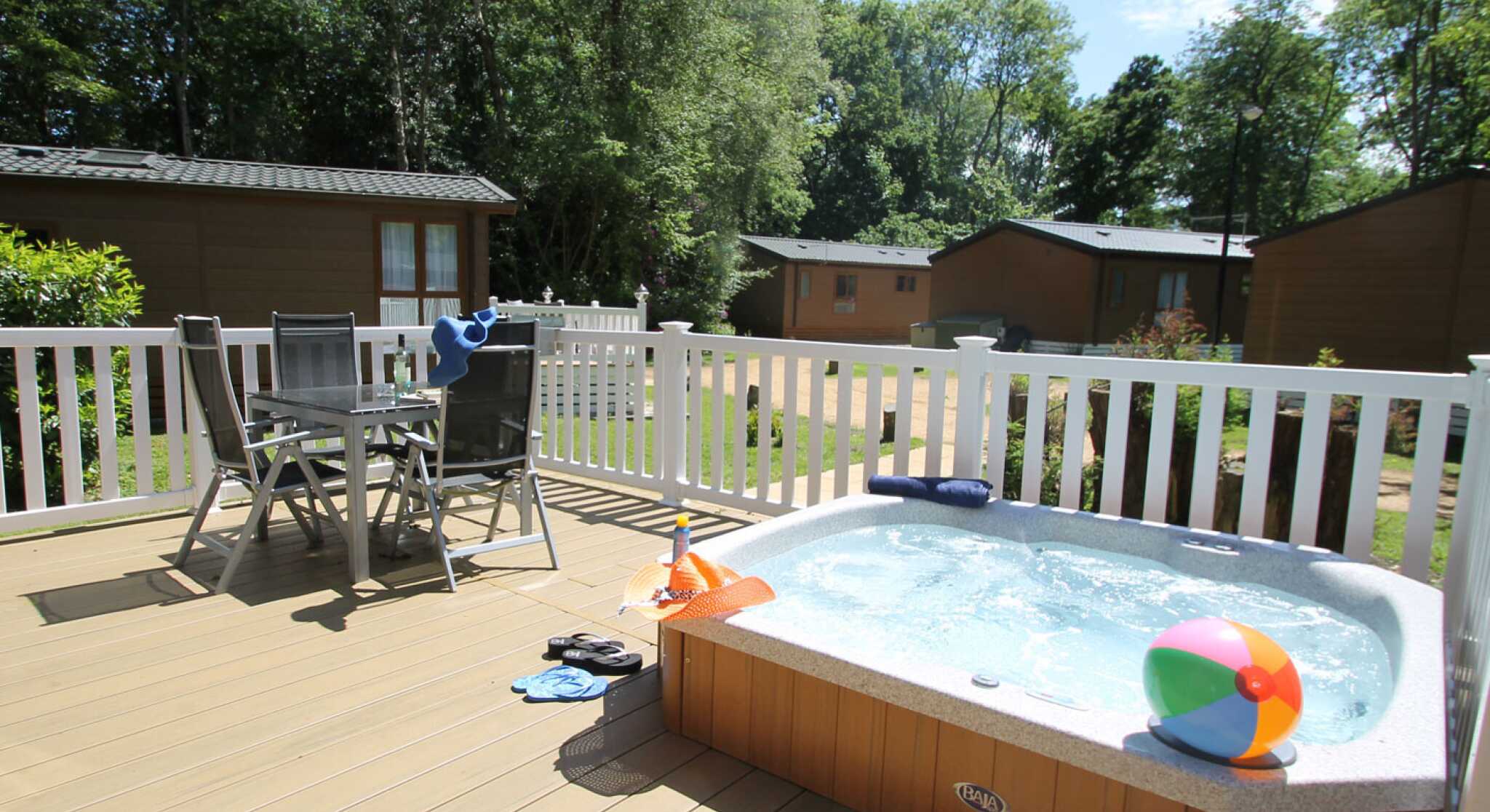 A hot tub on a wooden deck with a beach ball nearby. Two chairs and a small table are set up beside the tub, surrounded by greenery and cabins in the background. The sky is clear with a few clouds.