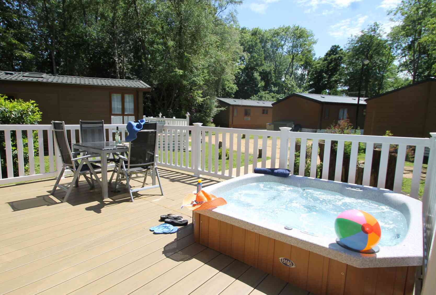 A hot tub on a wooden deck with a beach ball nearby. Two chairs and a small table are set up beside the tub, surrounded by greenery and cabins in the background. The sky is clear with a few clouds.