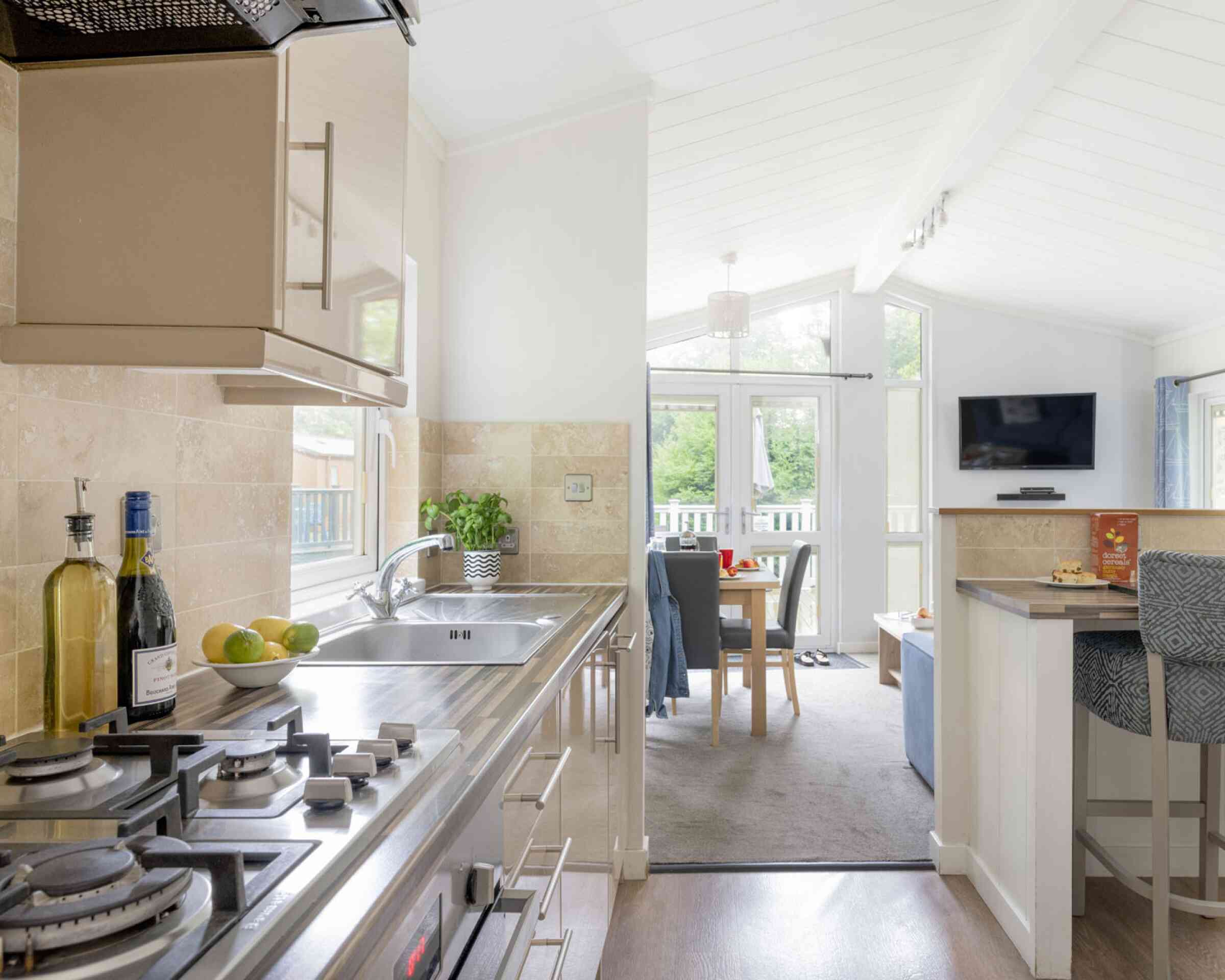Modern kitchen with stainless steel appliances and a gas stove. A light, airy dining area is visible in the background, featuring a table set for meals and a cozy seating area with a TV. Natural light floods in through large windows, enhancing the spacious atmosphere.