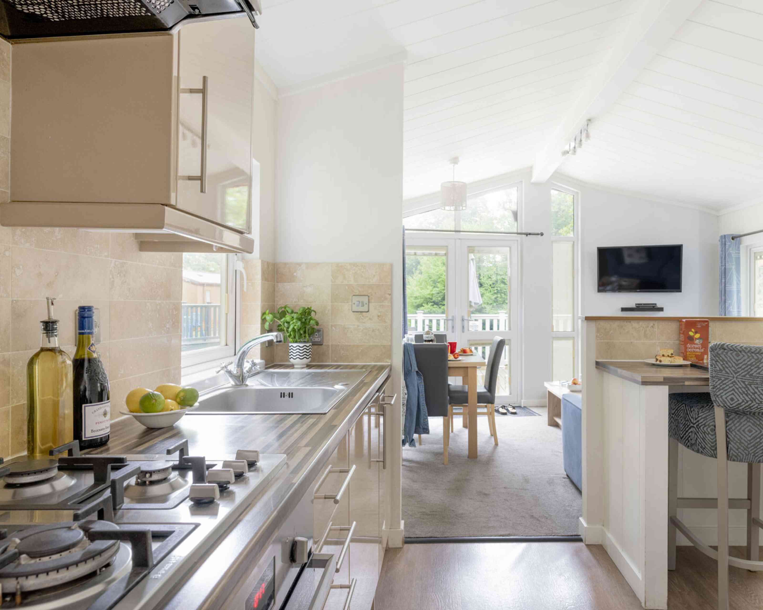 Modern kitchen with stainless steel appliances and a gas stove. A light, airy dining area is visible in the background, featuring a table set for meals and a cozy seating area with a TV. Natural light floods in through large windows, enhancing the spacious atmosphere.