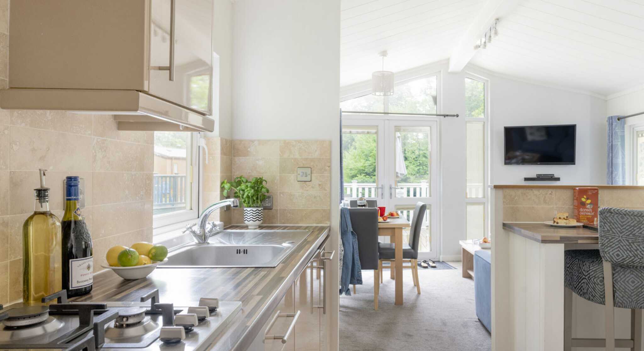 Modern kitchen with stainless steel appliances and a gas stove. A light, airy dining area is visible in the background, featuring a table set for meals and a cozy seating area with a TV. Natural light floods in through large windows, enhancing the spacious atmosphere.
