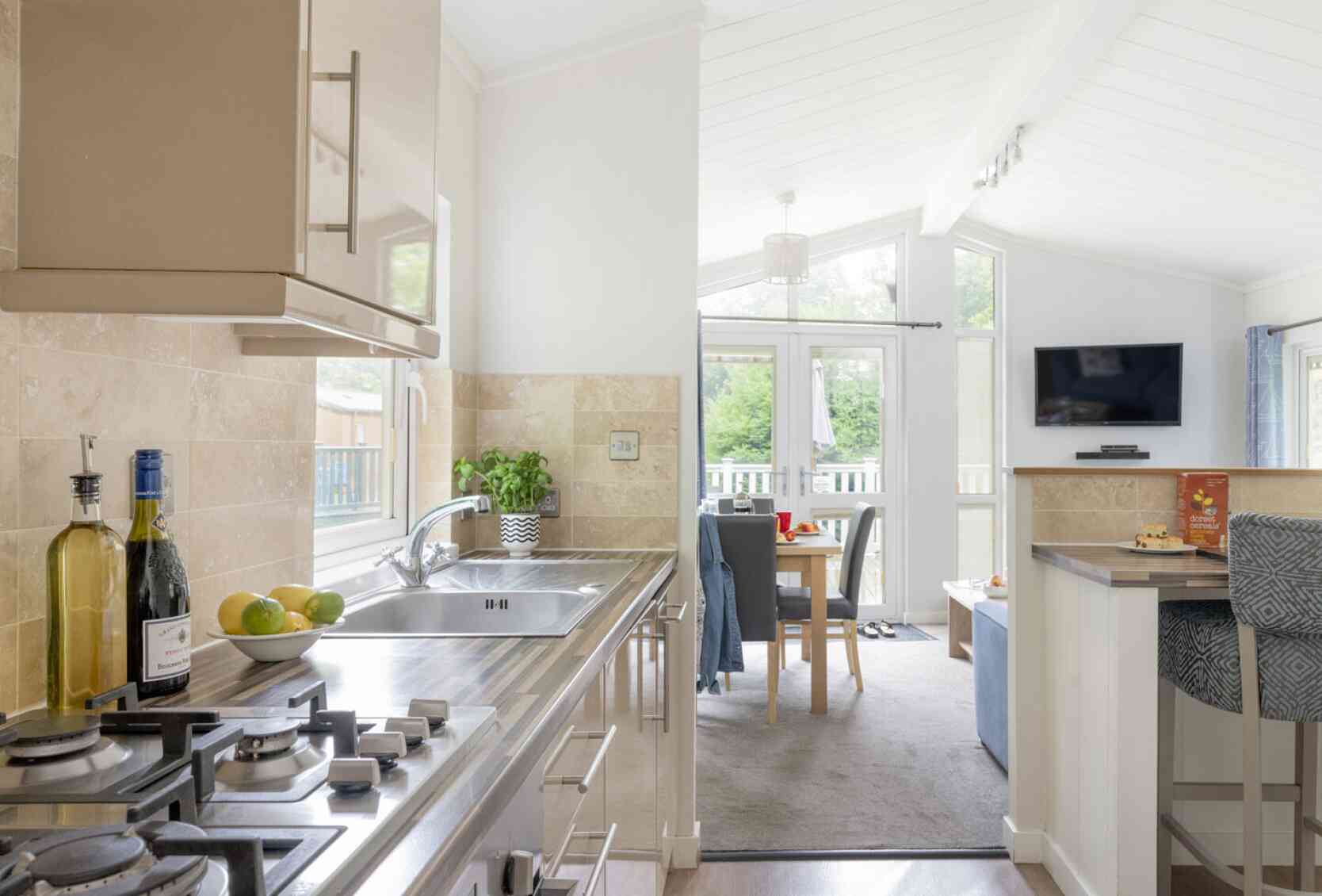 Modern kitchen with stainless steel appliances and a gas stove. A light, airy dining area is visible in the background, featuring a table set for meals and a cozy seating area with a TV. Natural light floods in through large windows, enhancing the spacious atmosphere.