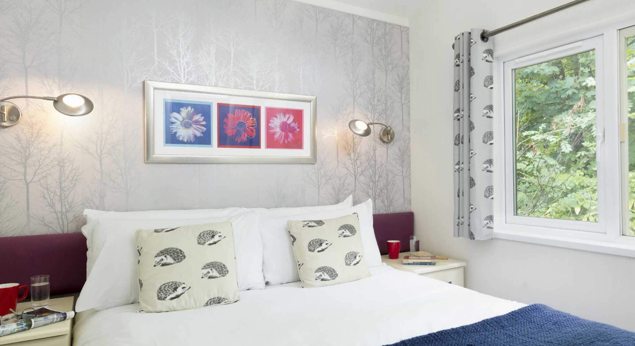 A cozy bedroom featuring a large bed with white linens and decorative pillows. A wall with floral artwork in red and blue complements light-colored walls adorned with a tree pattern. Two bedside tables with red cups sit on either side of the bed, and a window offers a view of greenery outside.