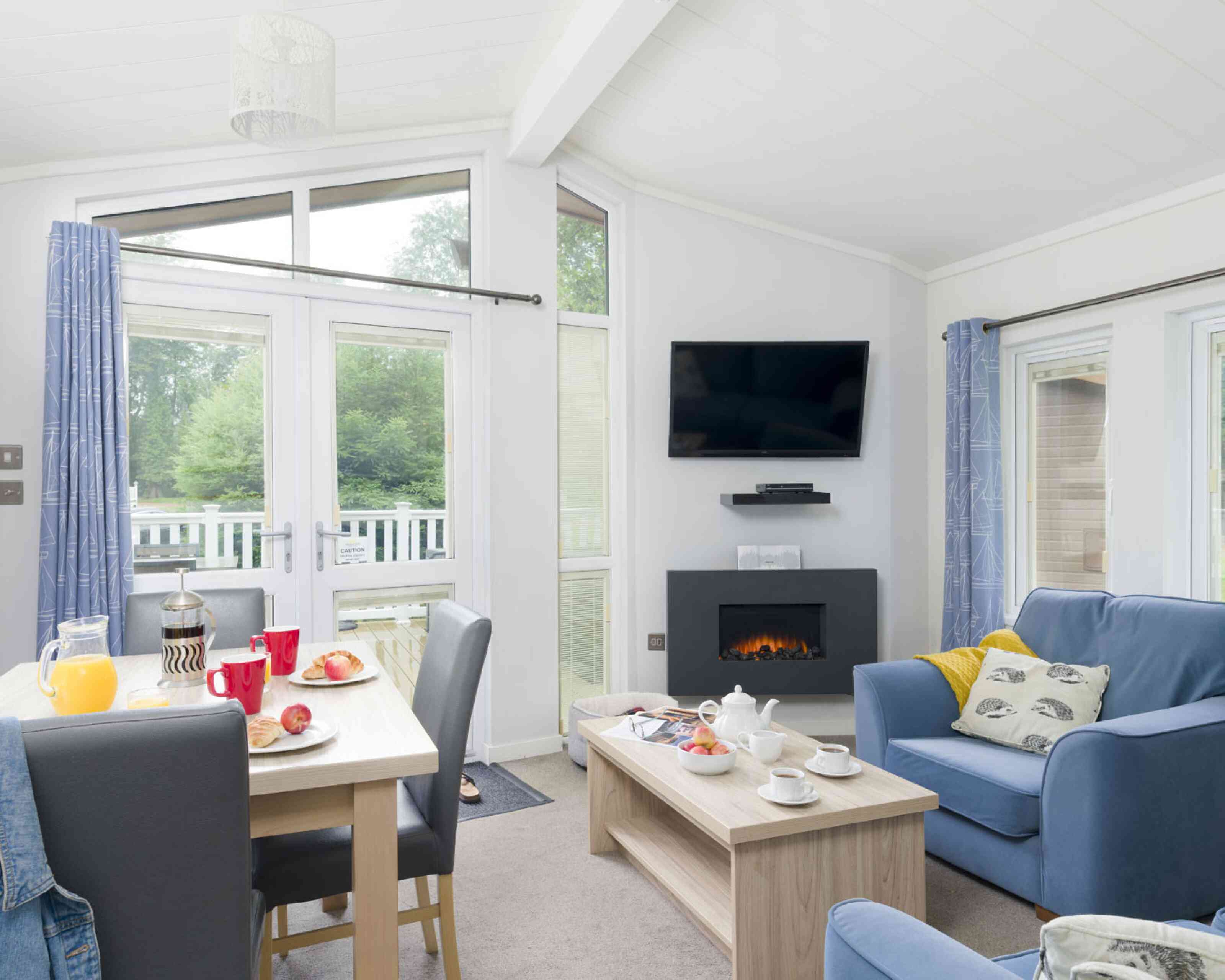 A cozy living space features a comfortable blue sofa, wooden coffee table, and a dining area with red mugs and a breakfast spread. A modern fireplace and a wall-mounted TV enhance the inviting atmosphere, with large windows allowing natural light and views of greenery outside.