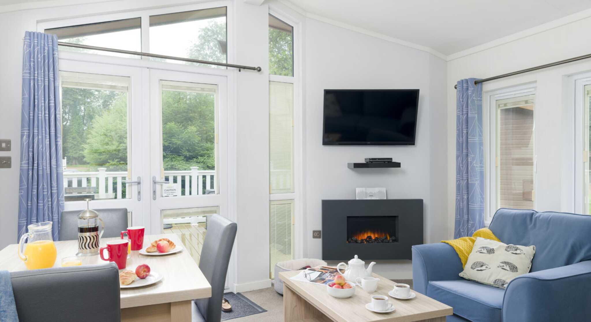 A cozy living space features a comfortable blue sofa, wooden coffee table, and a dining area with red mugs and a breakfast spread. A modern fireplace and a wall-mounted TV enhance the inviting atmosphere, with large windows allowing natural light and views of greenery outside.