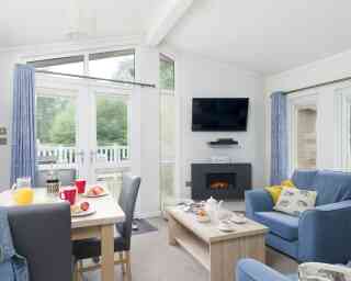 A cozy living space features a comfortable blue sofa, wooden coffee table, and a dining area with red mugs and a breakfast spread. A modern fireplace and a wall-mounted TV enhance the inviting atmosphere, with large windows allowing natural light and views of greenery outside.