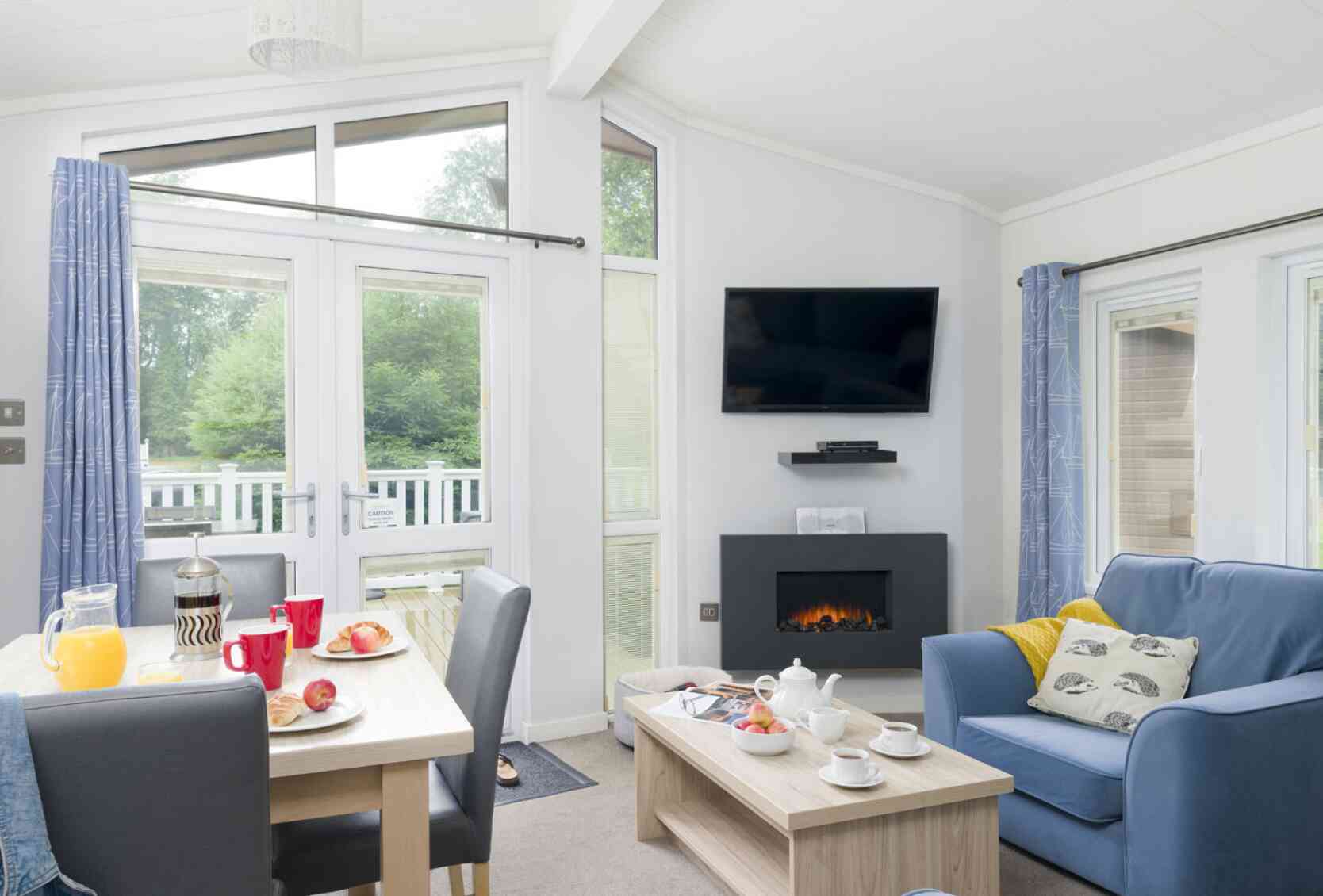 A cozy living space features a comfortable blue sofa, wooden coffee table, and a dining area with red mugs and a breakfast spread. A modern fireplace and a wall-mounted TV enhance the inviting atmosphere, with large windows allowing natural light and views of greenery outside.