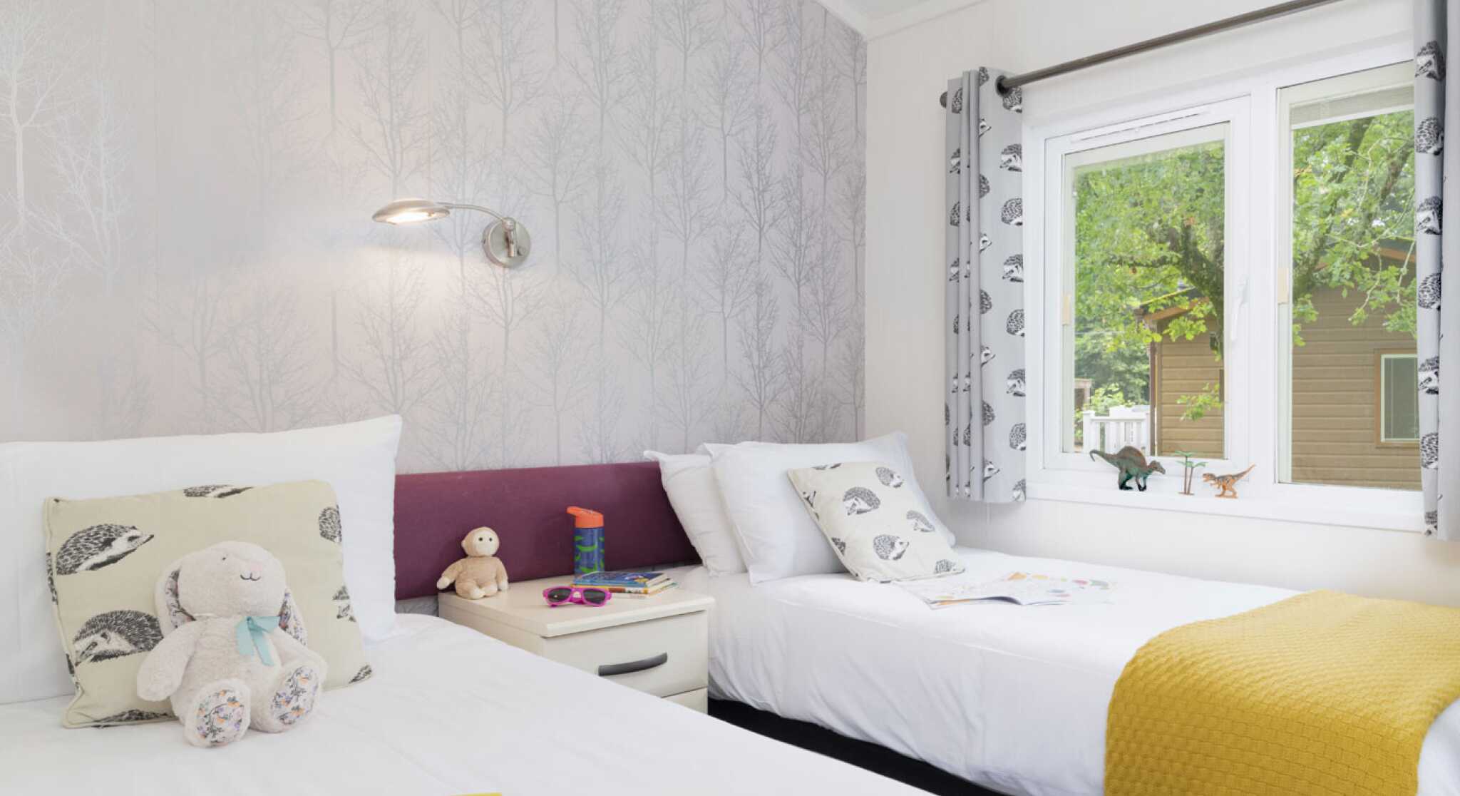 A bright and inviting bedroom featuring two twin beds, each with fluffy white bedding and decorative pillows. One bed has a yellow throw blanket, and a bedside table hosts a few children's books and a stuffed animal. The wall has a subtle tree-patterned wallpaper, and a large window lets in natural light, surrounded by patterned curtains.