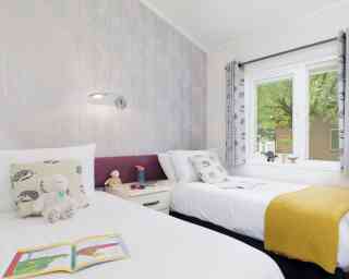 A bright and inviting bedroom featuring two twin beds, each with fluffy white bedding and decorative pillows. One bed has a yellow throw blanket, and a bedside table hosts a few children's books and a stuffed animal. The wall has a subtle tree-patterned wallpaper, and a large window lets in natural light, surrounded by patterned curtains.
