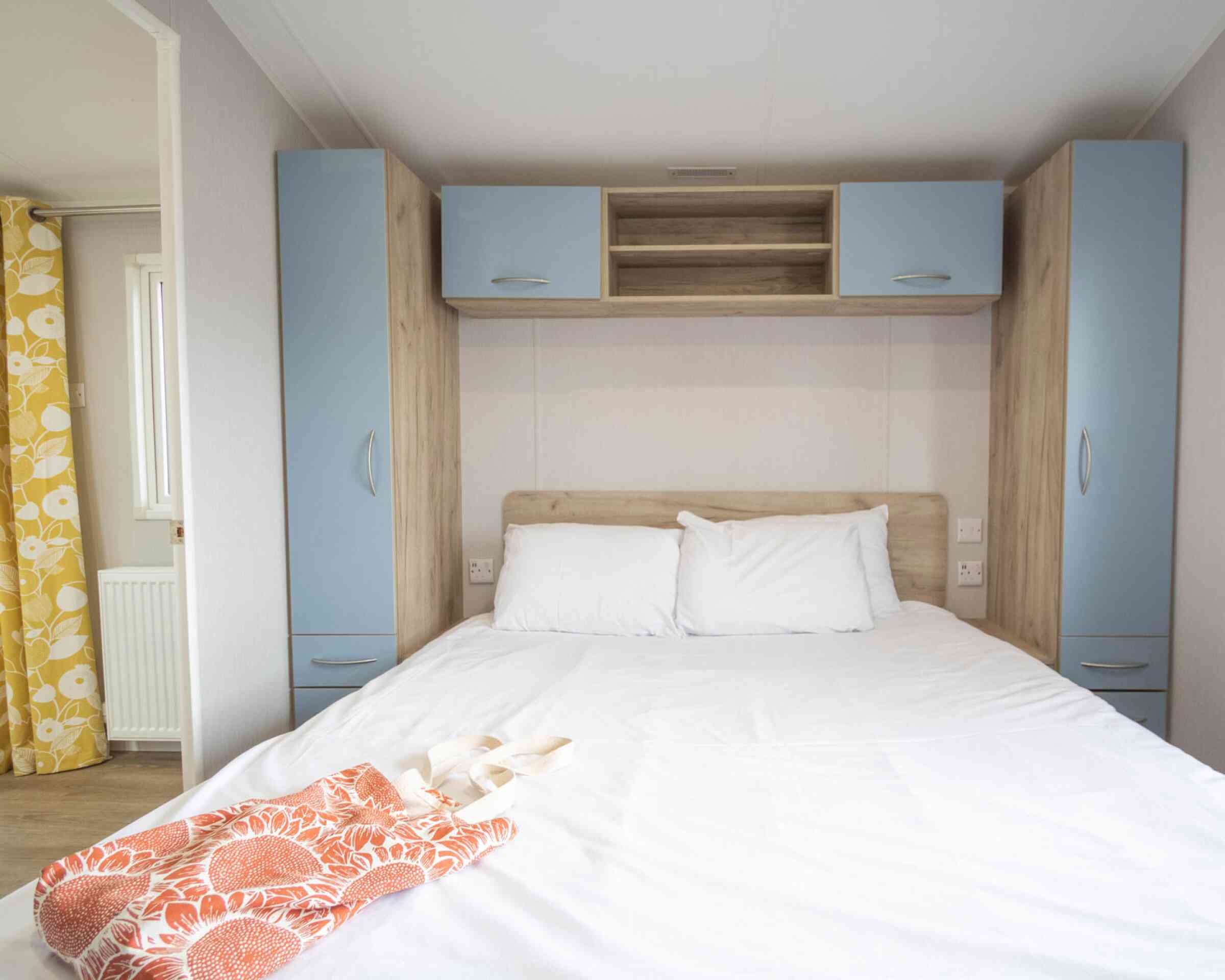 A cozy bedroom with a double bed featuring white bedding, light wooden furniture, and blue cabinets above. An orange bag rests on the bed, and a window with yellow patterned curtains is visible in the background.