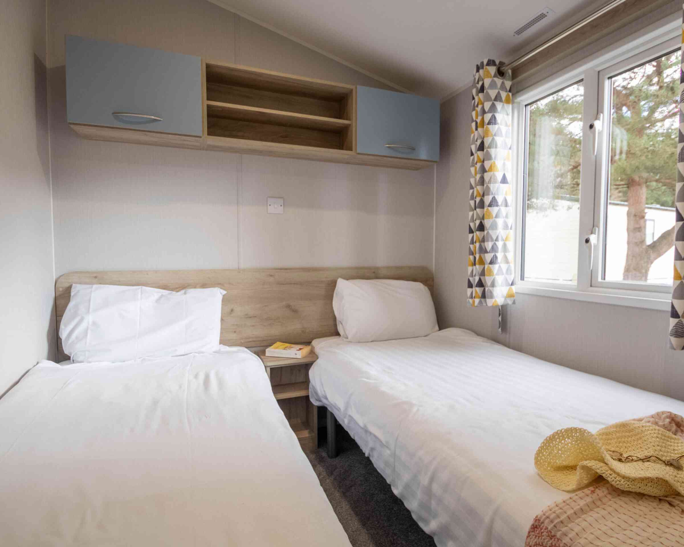 A cozy bedroom featuring two single beds with white linens, a wooden headboard, and a small shelf above. Large windows with patterned curtains allow natural light to brighten the room. A soft throw blanket is draped over one bed.