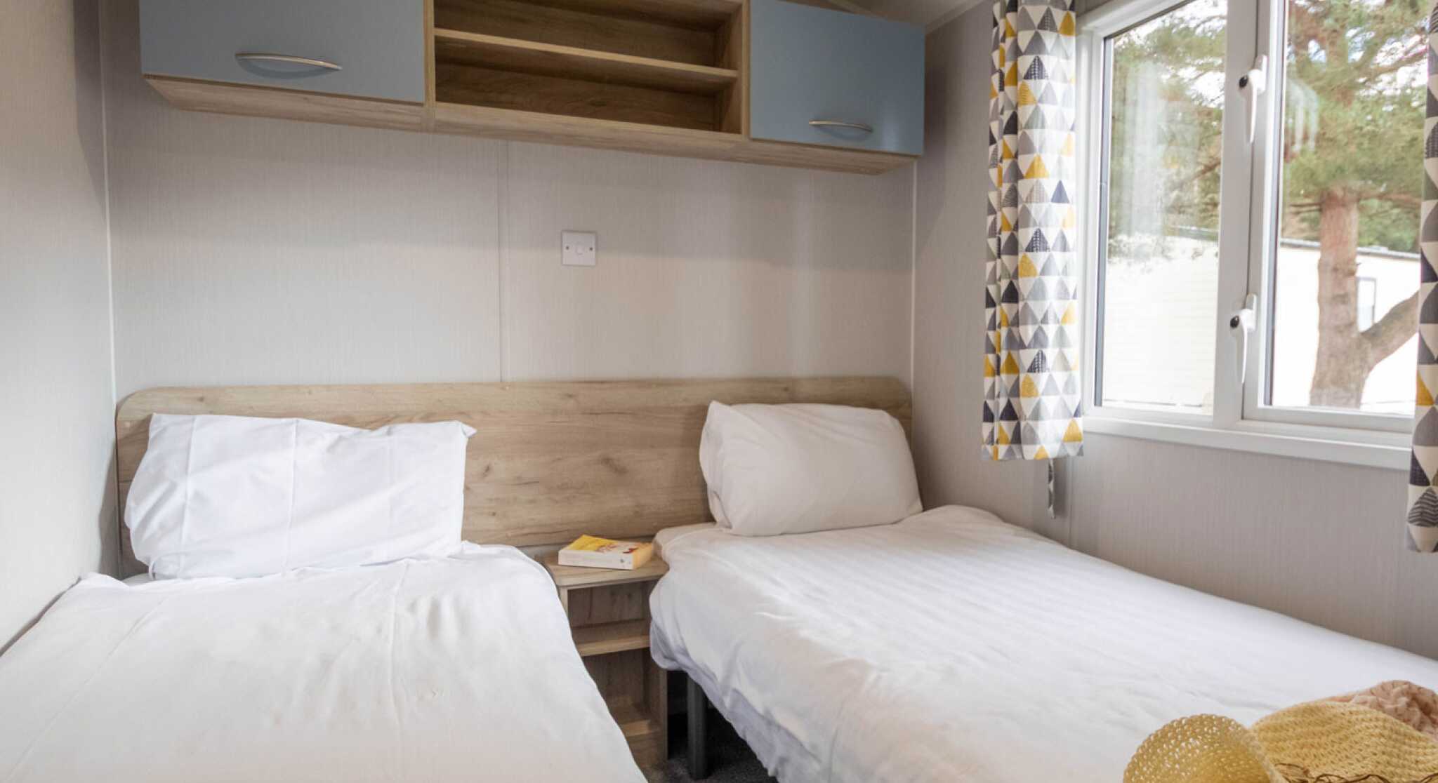 A cozy bedroom featuring two single beds with white linens, a wooden headboard, and a small shelf above. Large windows with patterned curtains allow natural light to brighten the room. A soft throw blanket is draped over one bed.