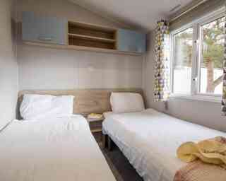 A cozy bedroom featuring two single beds with white linens, a wooden headboard, and a small shelf above. Large windows with patterned curtains allow natural light to brighten the room. A soft throw blanket is draped over one bed.