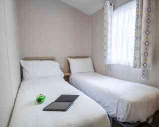 A small, cozy bedroom featuring two single beds with white bedding. A green and black toy sits on one bed, while an open notebook lies on the other. Natural light streams through a window dressed with patterned curtains.