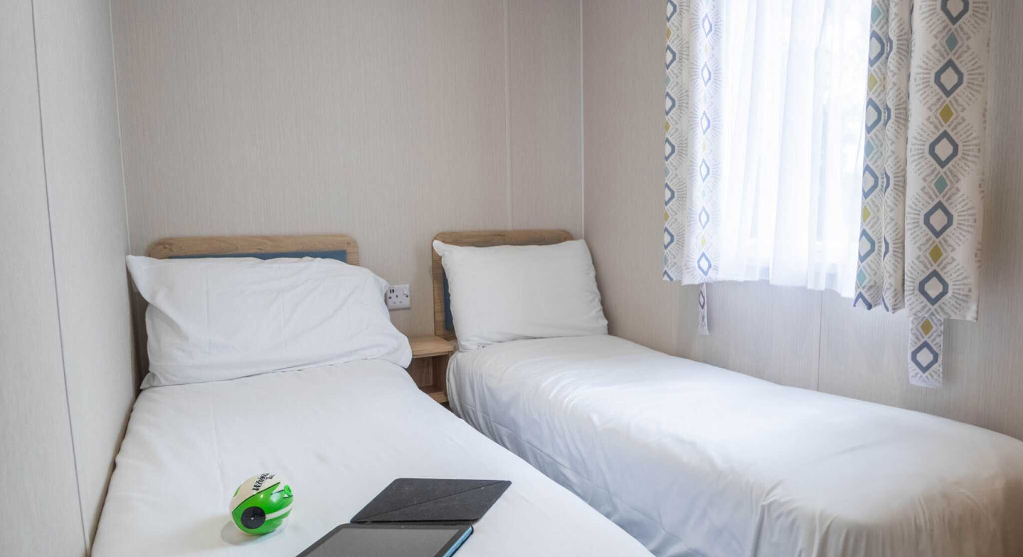 A small, cozy bedroom featuring two single beds with white bedding. A green and black toy sits on one bed, while an open notebook lies on the other. Natural light streams through a window dressed with patterned curtains.