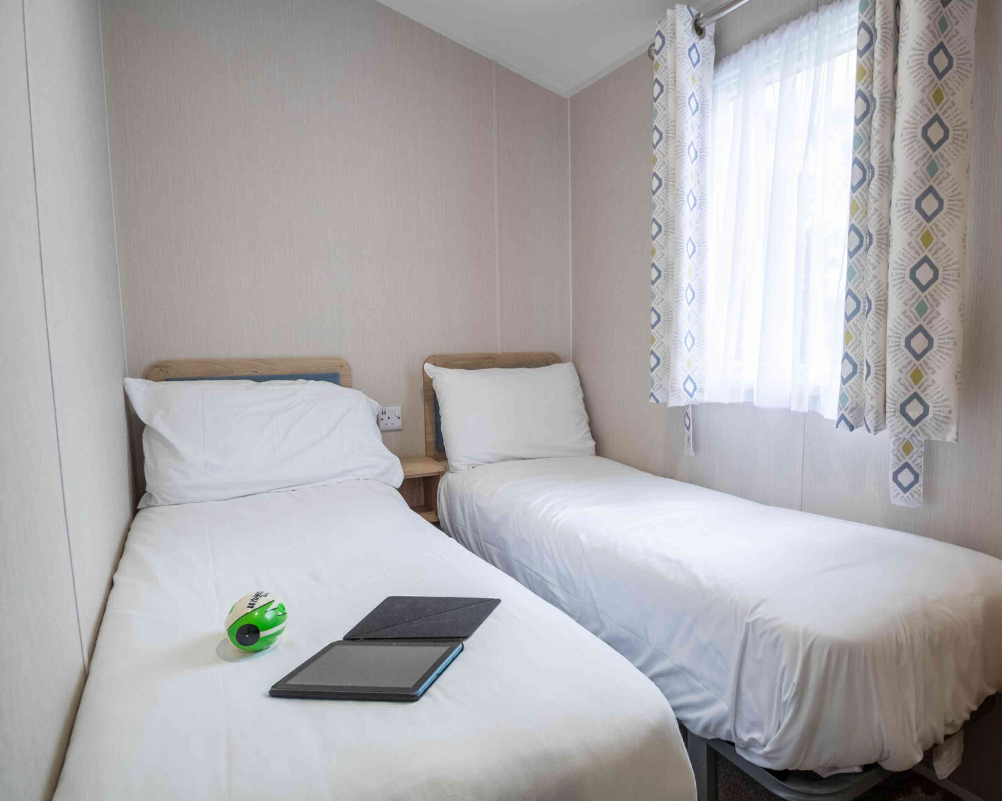 A small, cozy bedroom featuring two single beds with white bedding. A green and black toy sits on one bed, while an open notebook lies on the other. Natural light streams through a window dressed with patterned curtains.