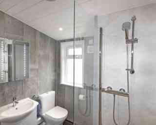 Modern bathroom featuring a glass shower enclosure, a toilet, and a vanity with a sink. The walls are tiled in a light gray, and a backlit mirror is mounted above the sink. Natural light enters through a window.