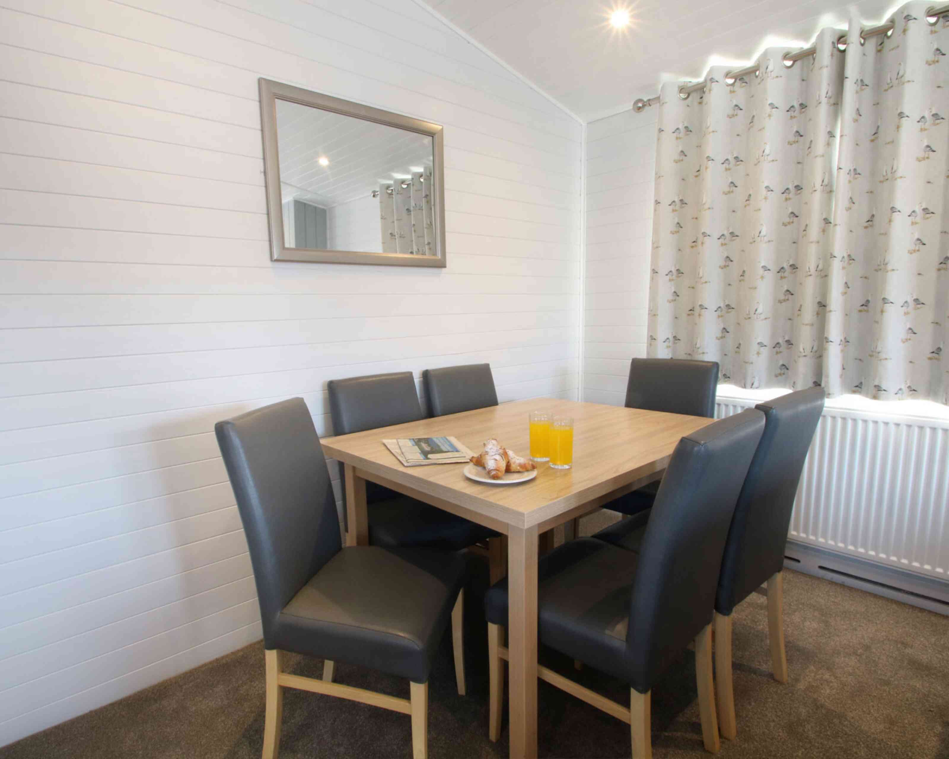 A cozy dining area with a wooden table surrounded by six black leather chairs. A plate with pastries and two glasses of orange juice sit on the table. A mirror hangs on the wall, reflecting the soft light from a nearby window, which is dressed with patterned curtains.